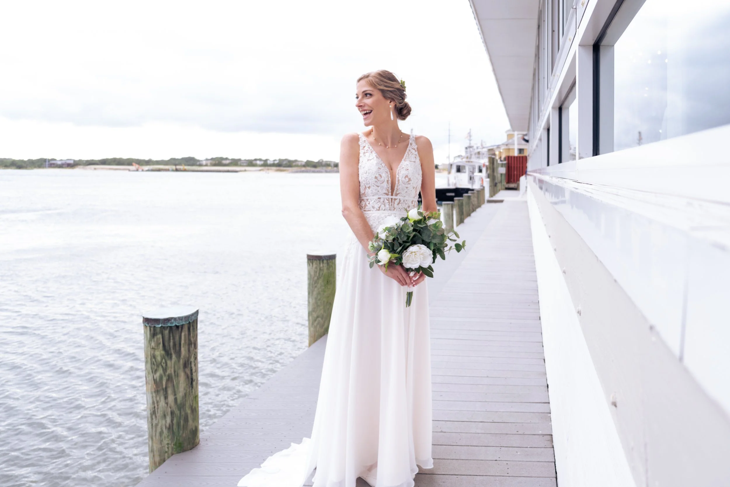 A smiling bride in a white lace wedding dress holding a bouquet of white roses and greenery, standing on a dock beside water with cloudy sky in the background.