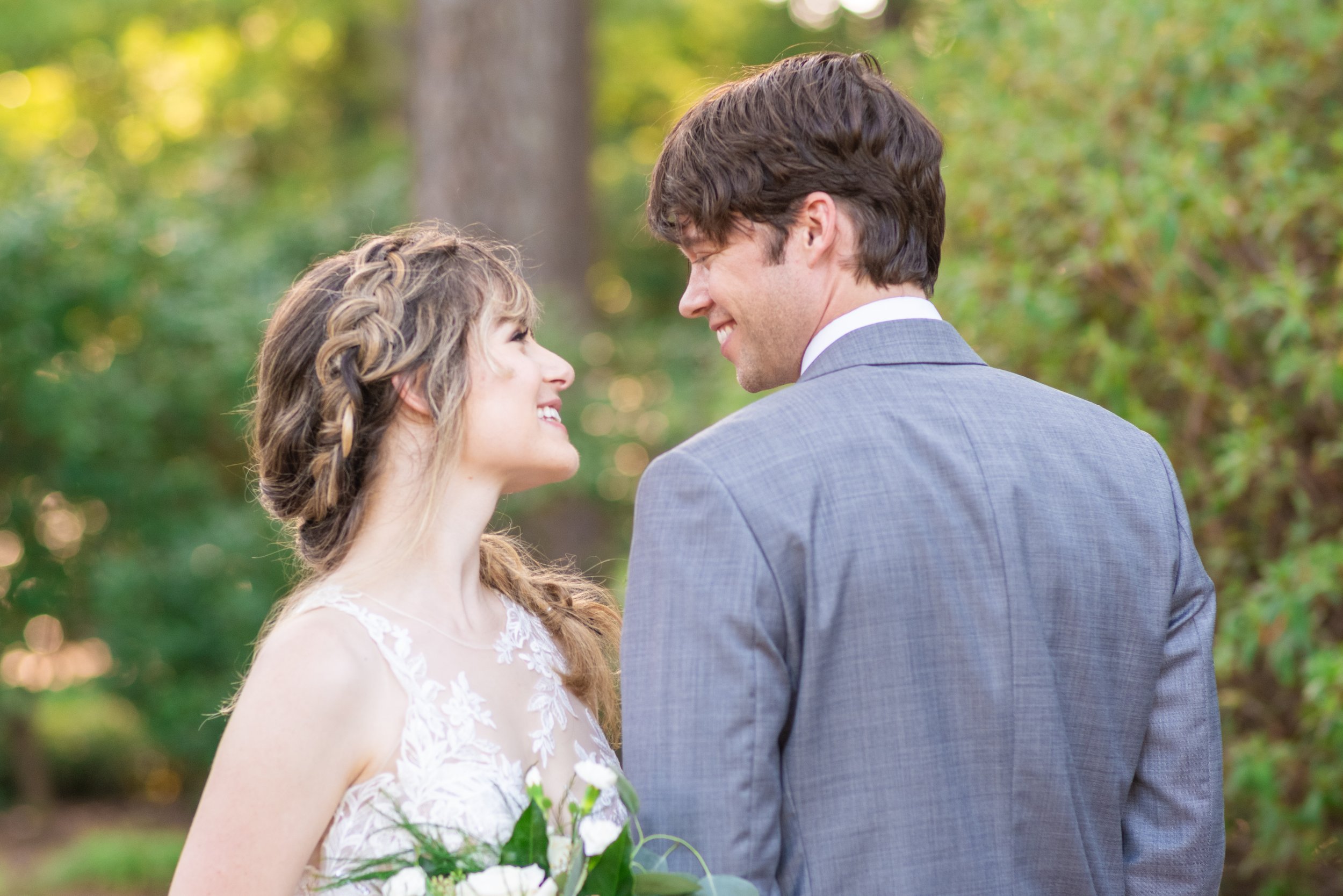 A smiling bride in a white lace wedding dress and a groom in a gray suit stand close together outdoors, gazing into each other's eyes amidst green trees and sunlight.