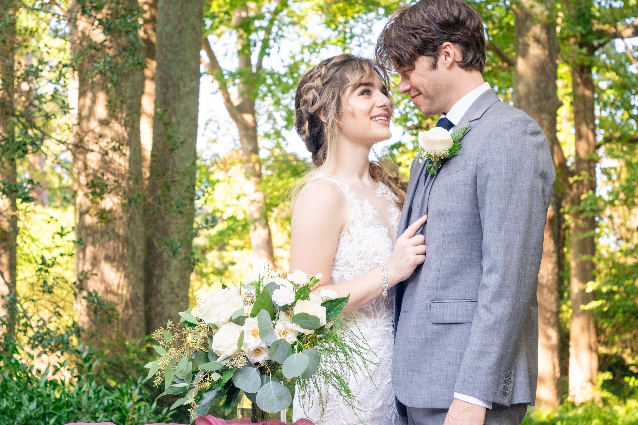 A bride and groom look at each other happily during their outdoor wedding ceremony in a wooded area, with the bride holding a bouquet of white flowers and green foliage.