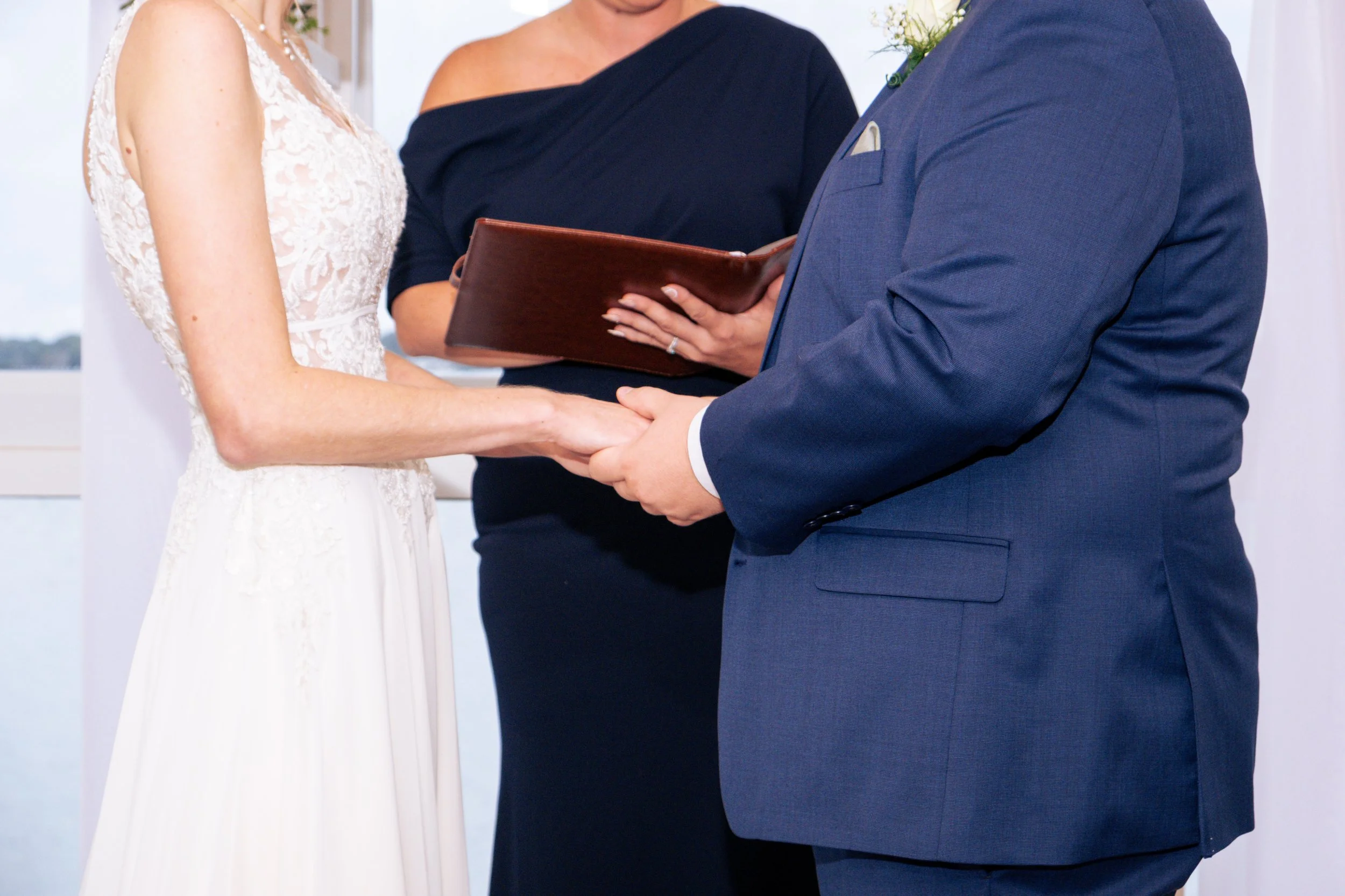 A wedding ceremony where a bride and groom are exchanging vows, holding hands, with a officiant present. The bride is wearing a lace wedding dress, and the groom is in a blue suit. The officiant is holding a leather folder.