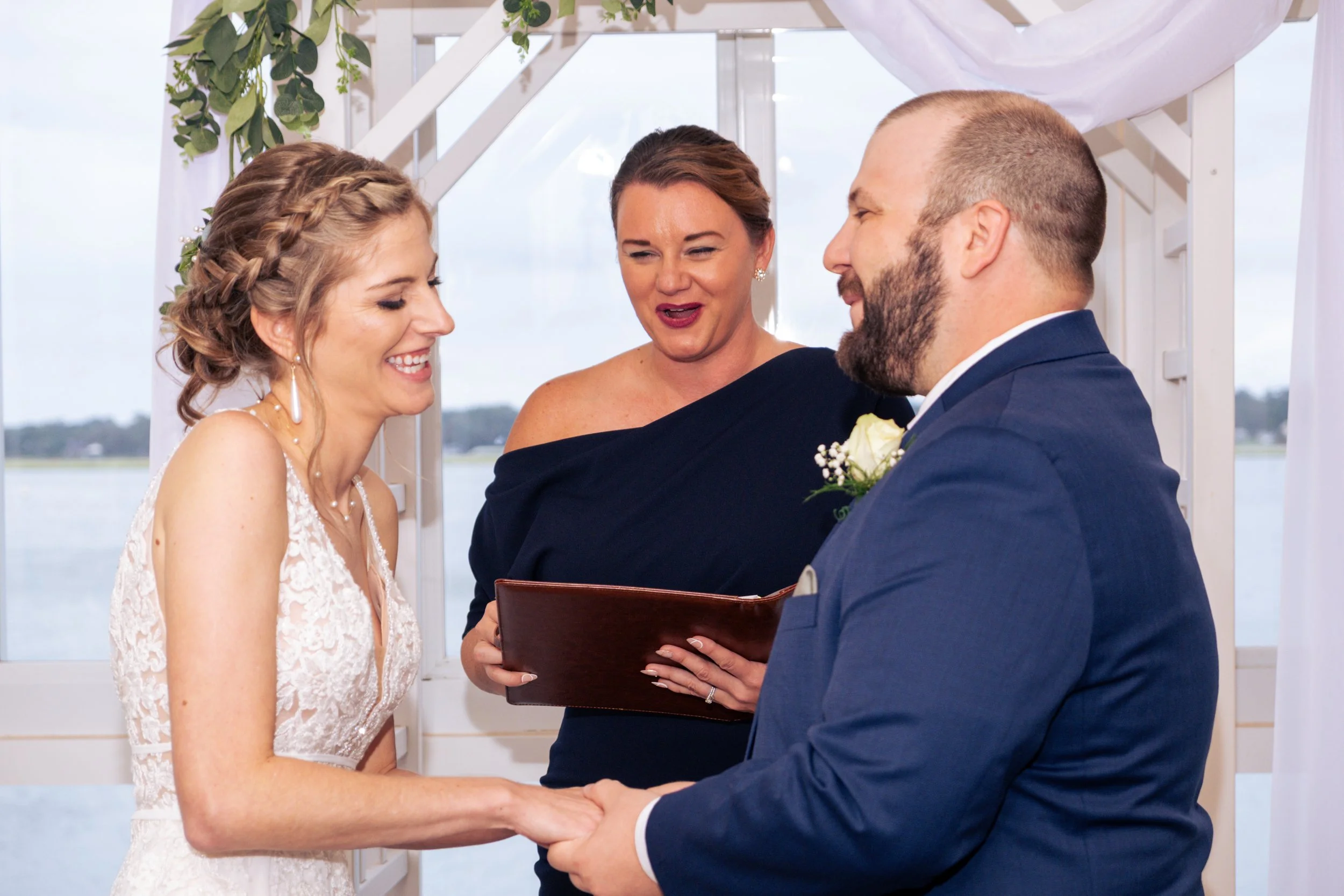 A bride and groom exchanging vows during their wedding ceremony, with an officiant holding a book standing behind them. The bride has braided hair and is wearing a lace dress, while the groom is in a dark suit with a white shirt, holding the bride's 