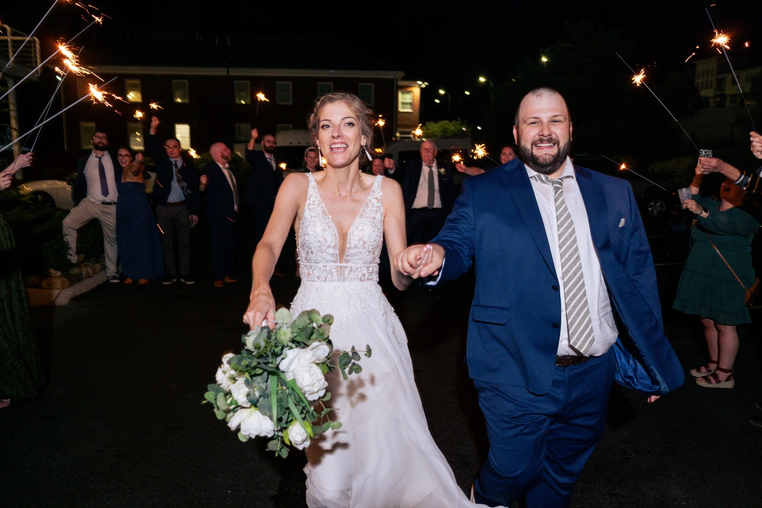 A newlywed couple walks outdoors at night, holding hands and smiling, surrounded by friends and family holding sparklers during a celebration.