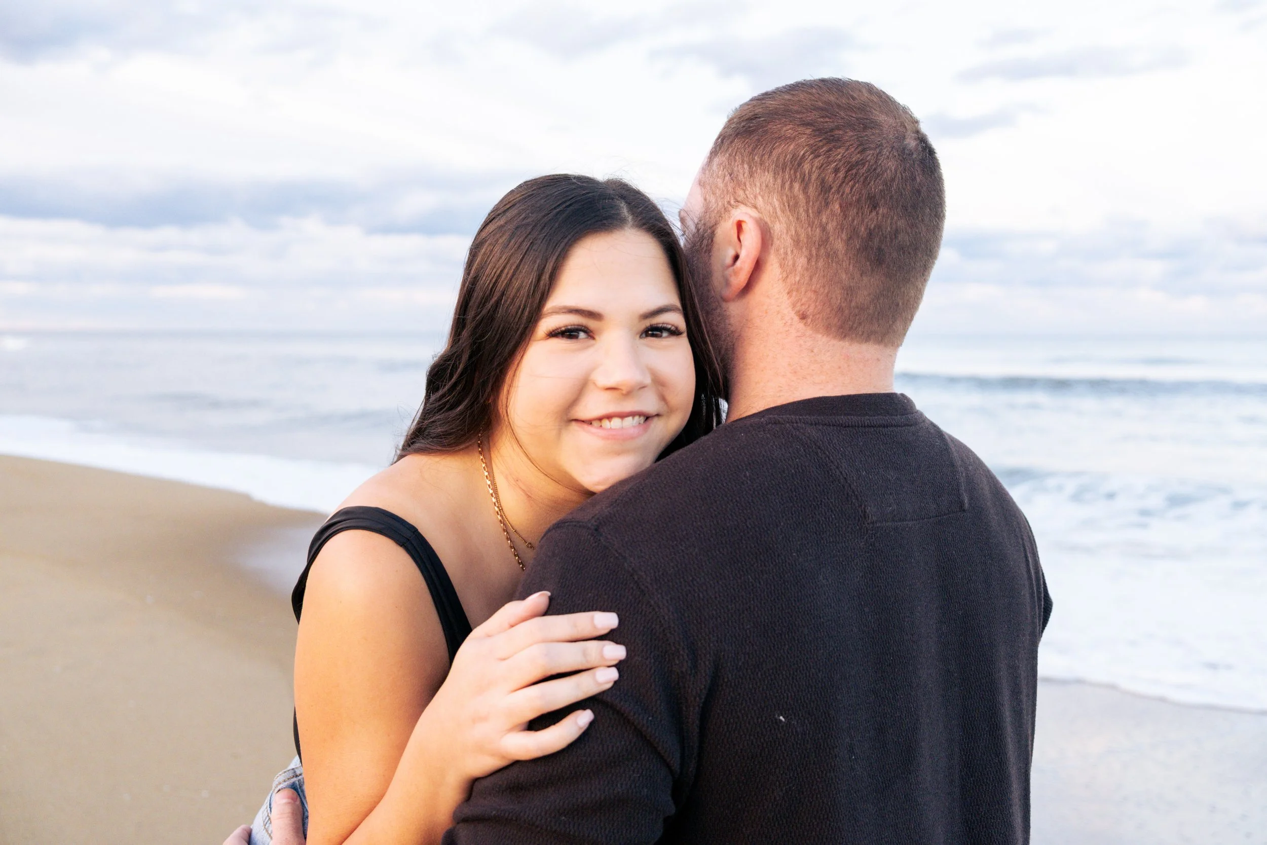 A young woman with long dark hair smiling while hugging a man with short light hair at the beach with ocean waves and cloudy sky in the background.