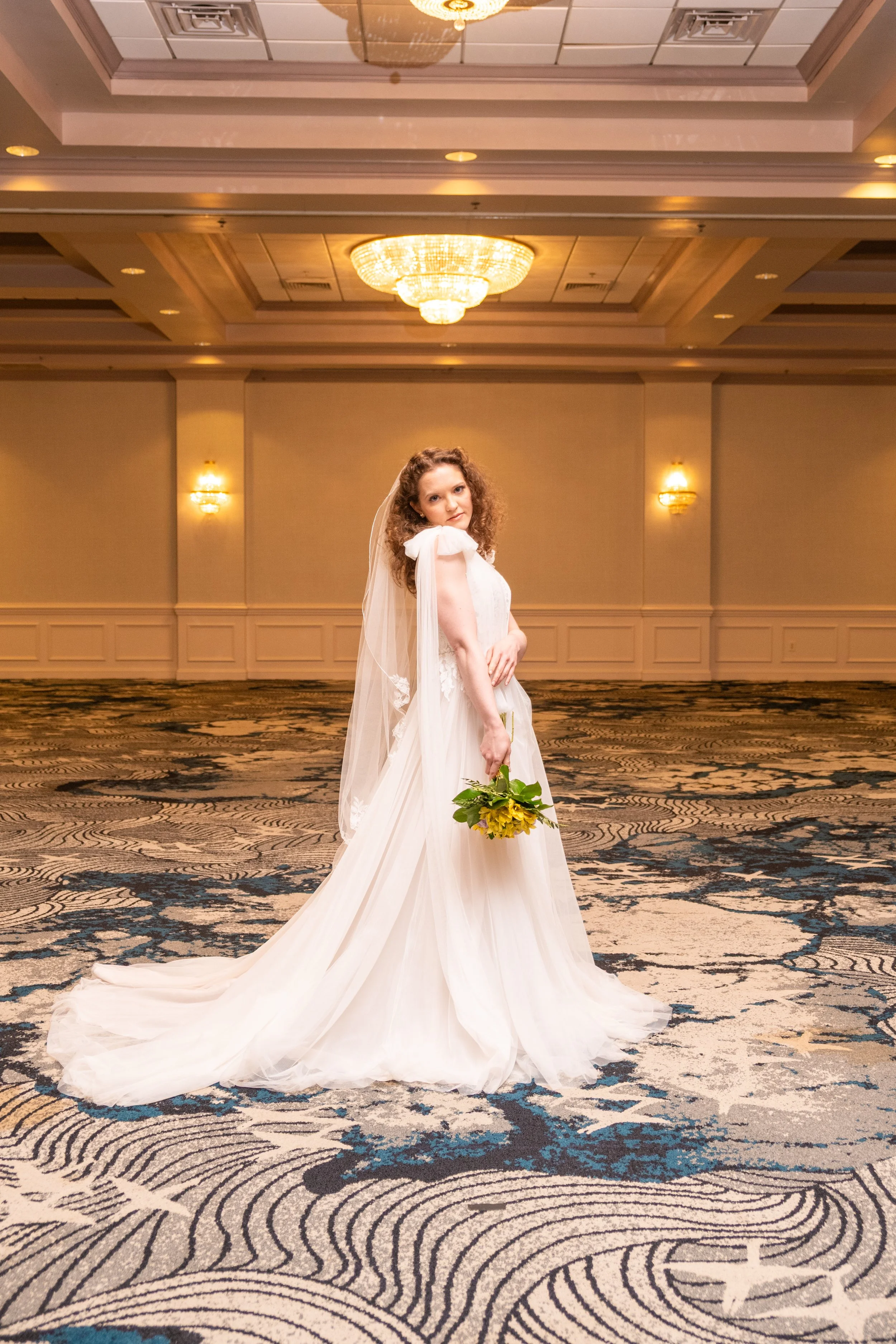 A bride with curly hair in a white wedding dress holding a bouquet of yellow flowers standing in a large, elegant banquet hall with chandeliers and patterned carpet.