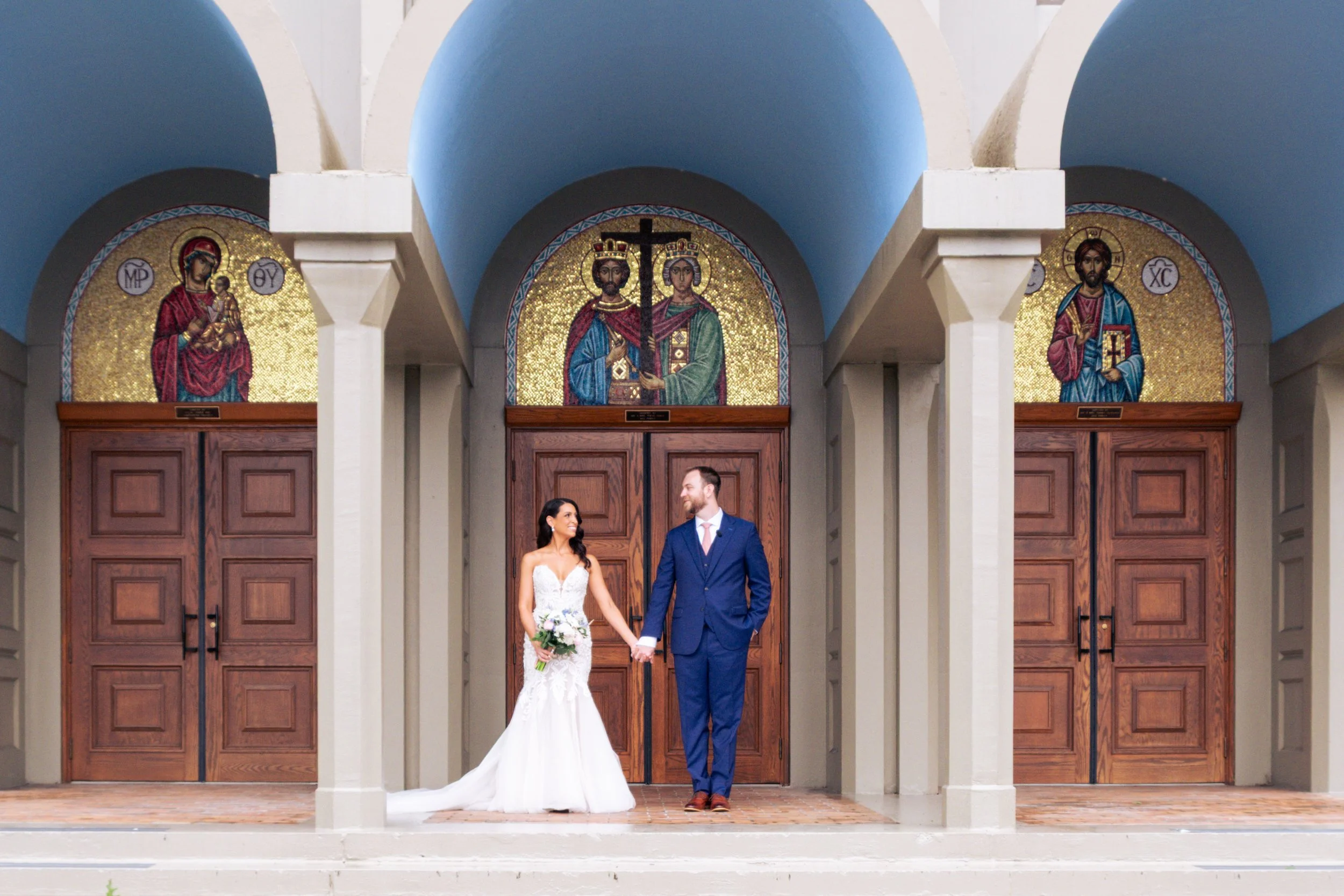 A bride and groom holding hands, standing in front of church doors, with religious icons above them, celebrating their wedding.