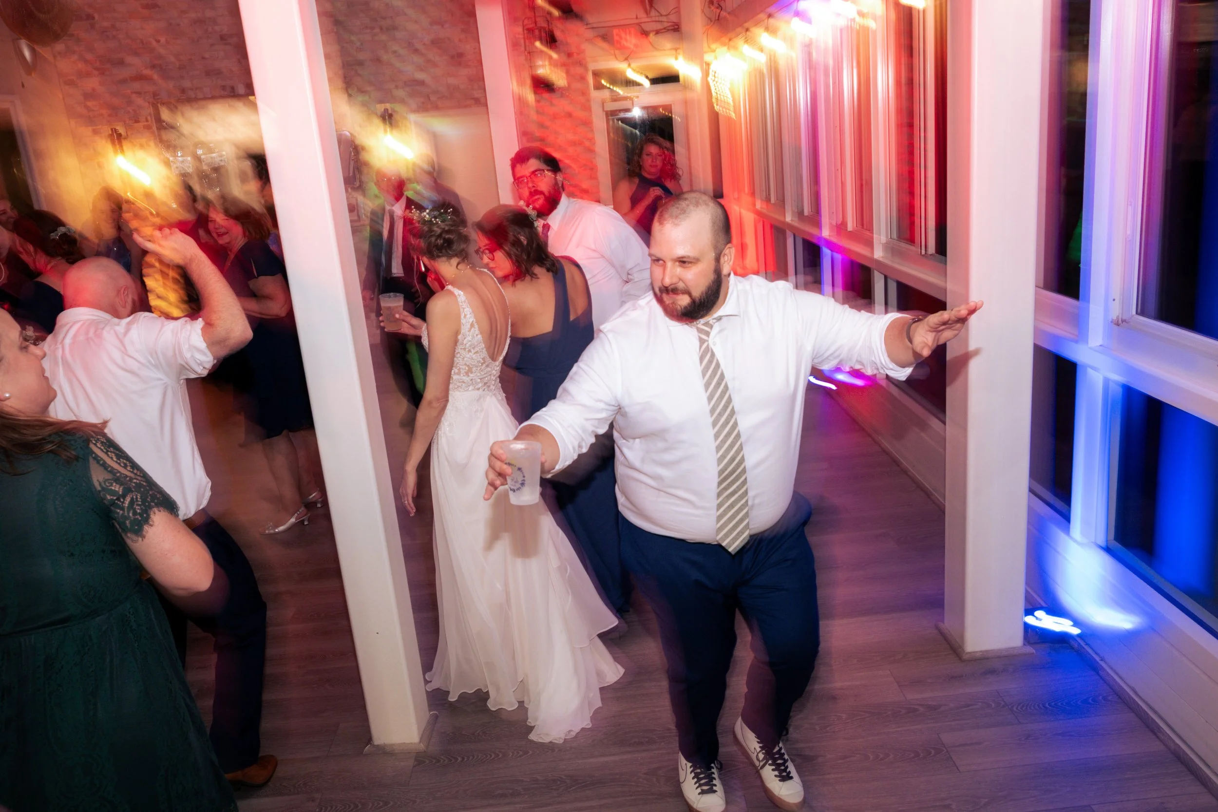 People dancing and socializing at a lively indoor celebration with colorful lights, a brick wall, and large windows.