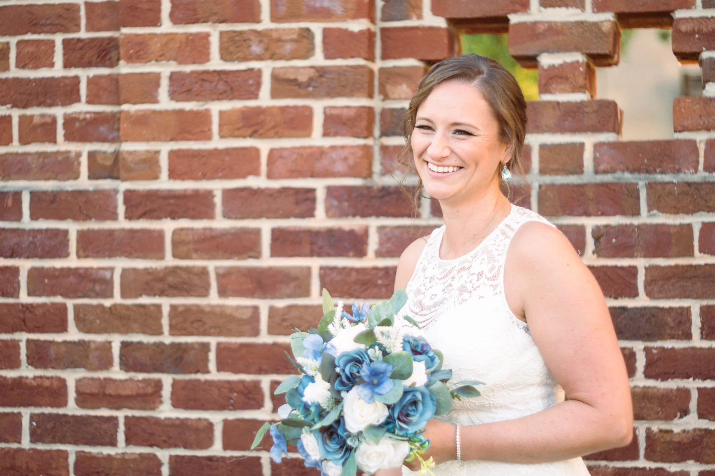 A woman in a white lace dress holding a bouquet of white and blue flowers, standing in front of a red brick wall, smiling and winking.