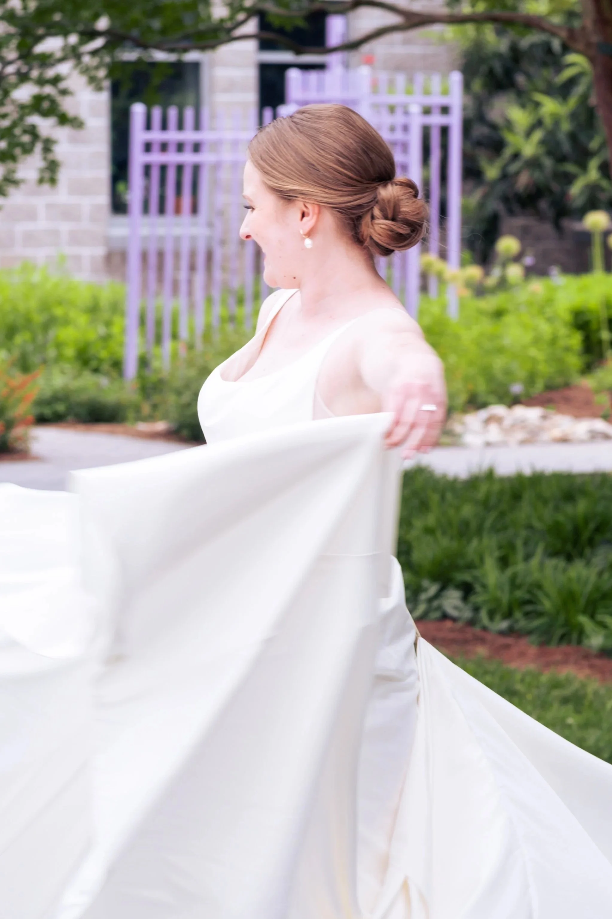 Side view of a woman with brown hair styled in a bun, wearing pearl earrings and a white dress, outdoors in a garden with green plants and a purple fence in the background.