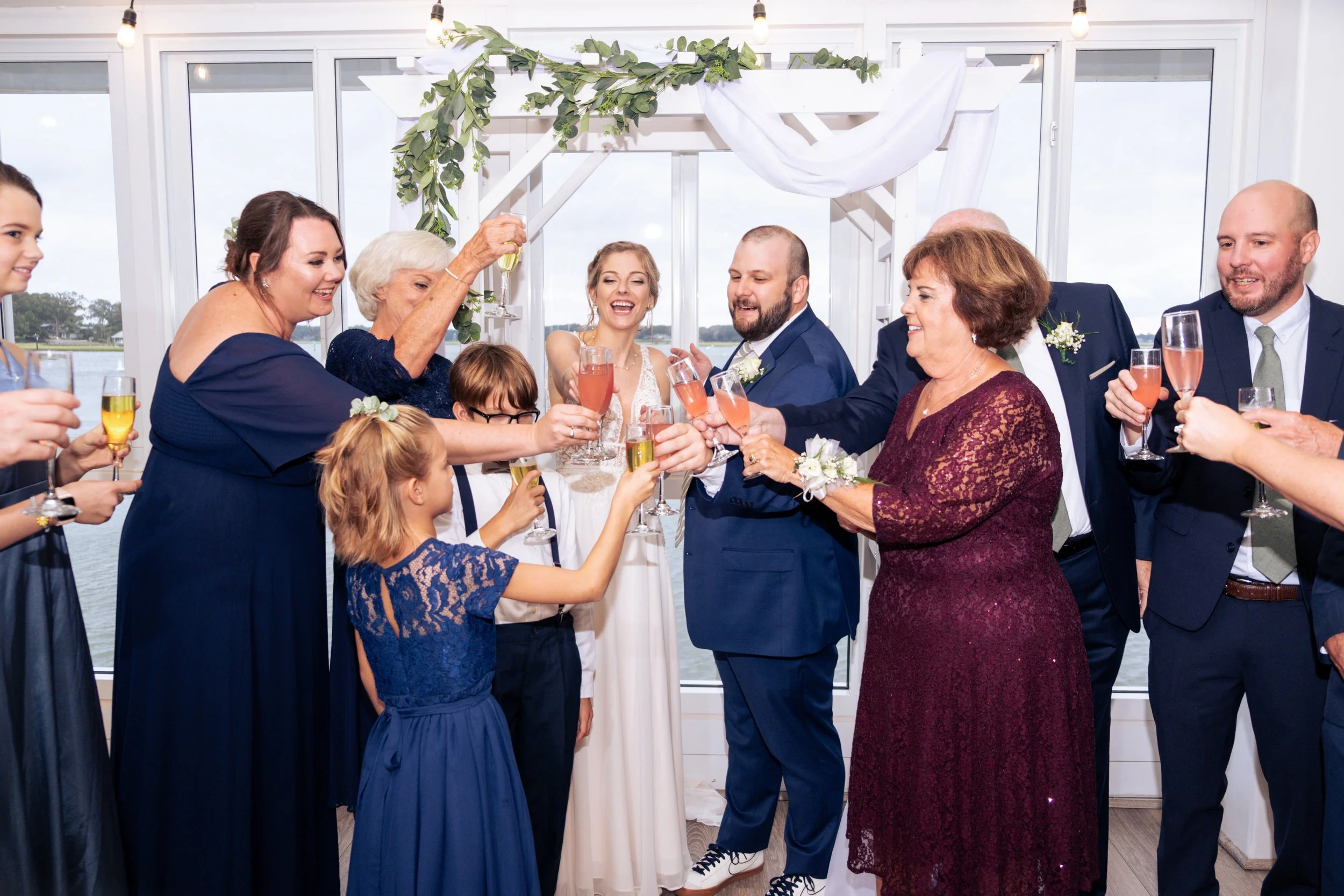 Group of people celebrating at a wedding reception, raising champagne glasses in a toast, with the bride and groom in the center, inside a decorated venue with large windows and greenery.
