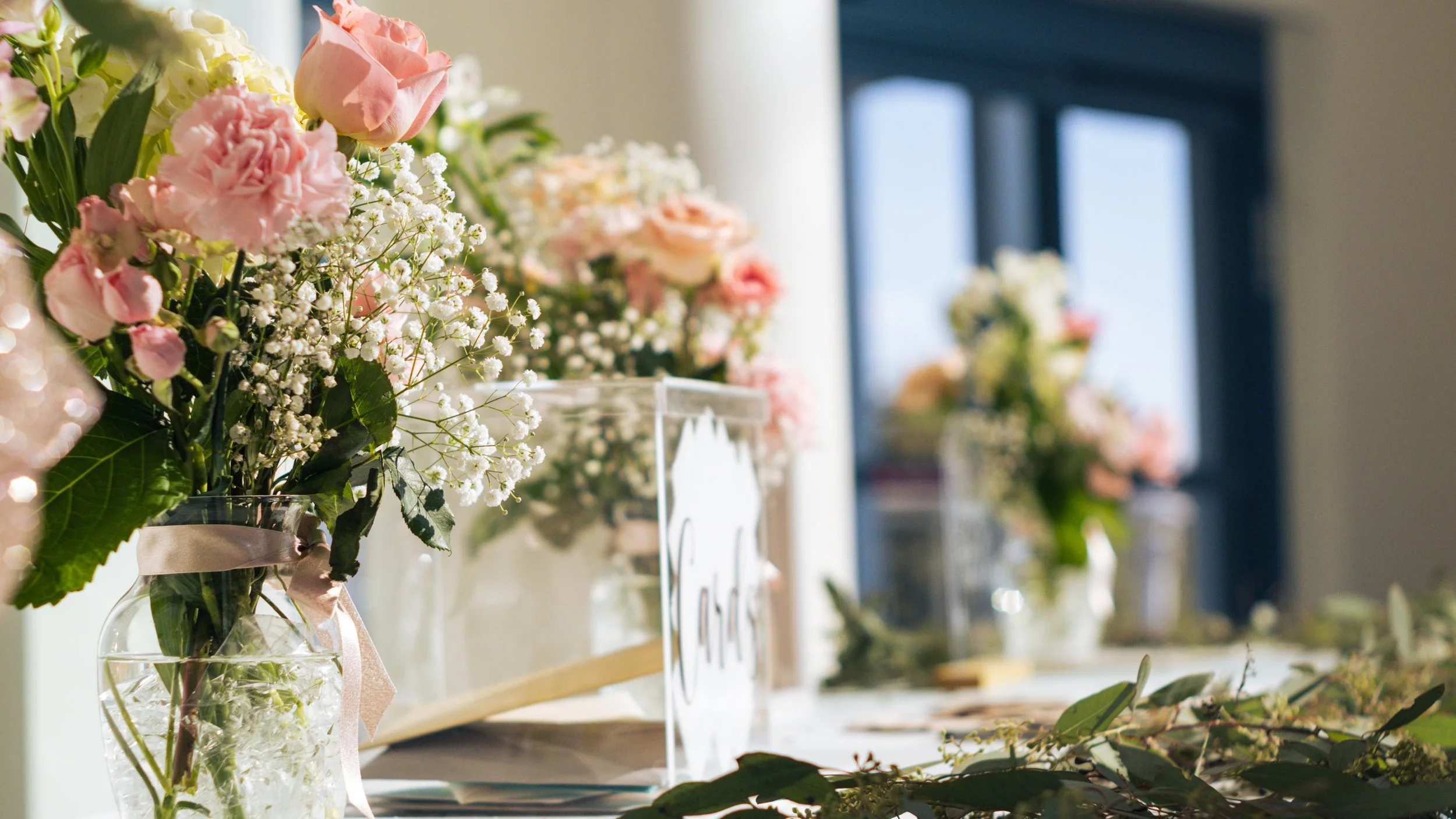 Pink and white flowers arranged in a glass vase on a table, with other similar arrangements in the background, possibly at a floral or event display.