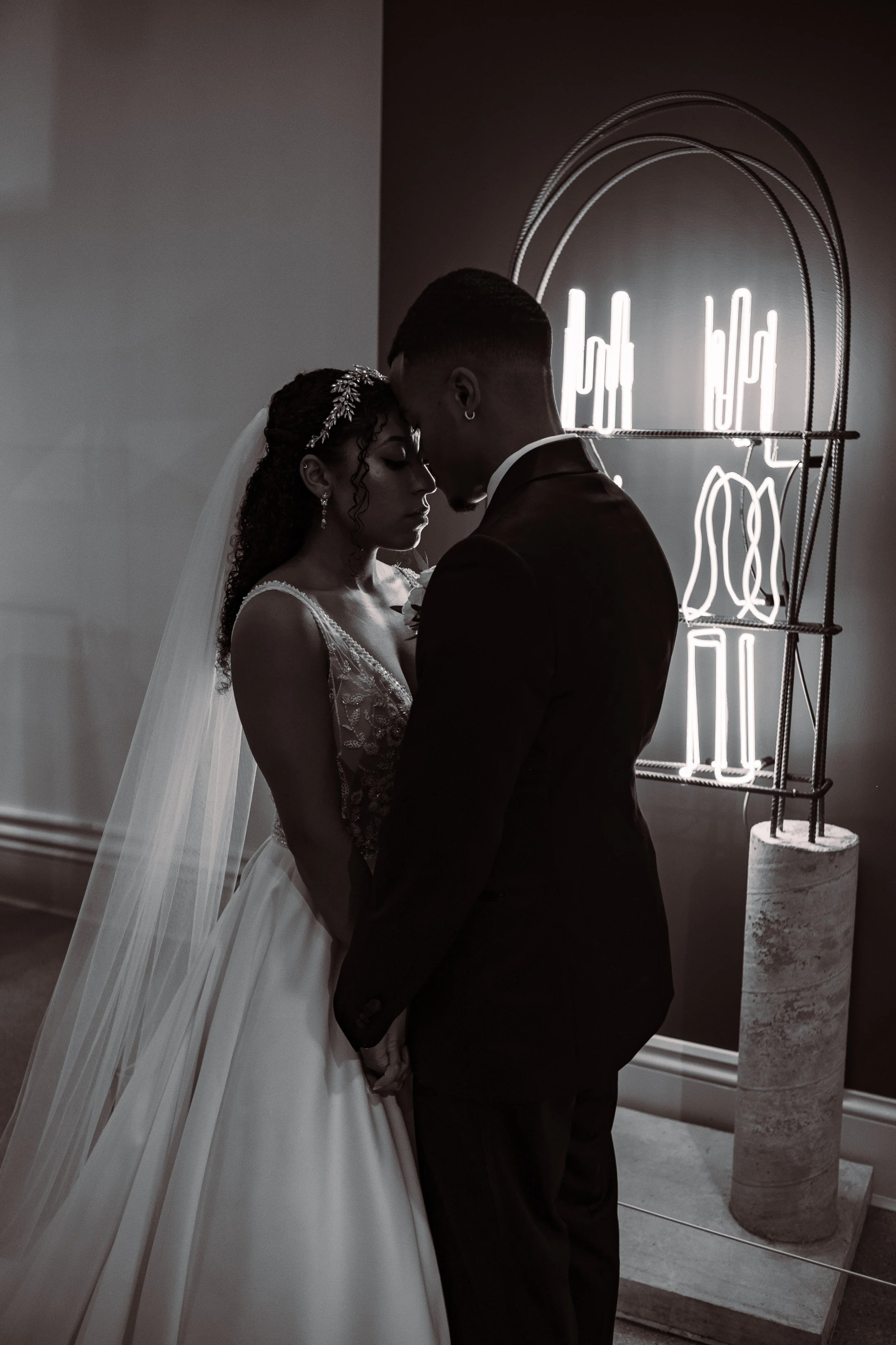 A wedding couple stands close, touching foreheads, with a neon light display in the background.