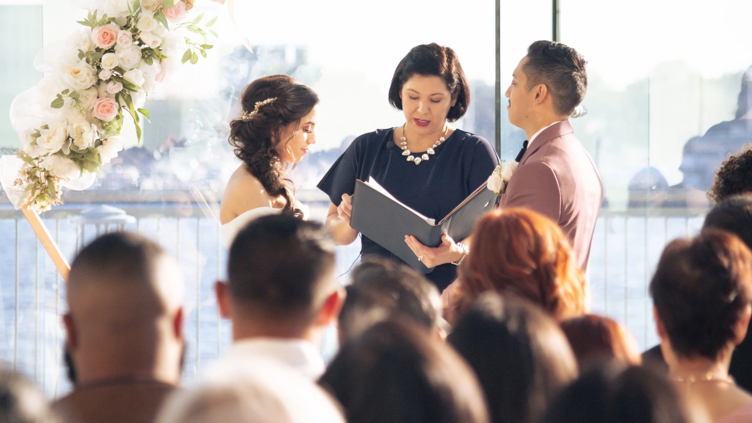 A wedding ceremony with a bride and groom standing before a officiant, with a decorated floral arch, in a bright venue with glass windows.