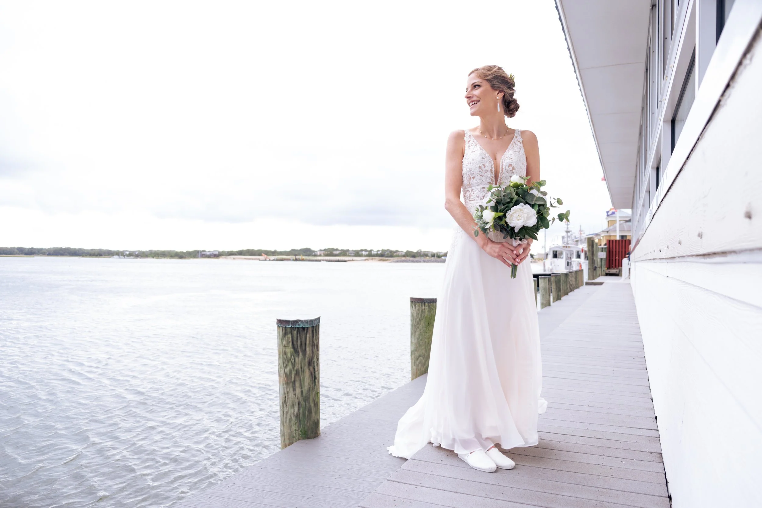 A bride in a white lace wedding dress holding a bouquet of white roses and greenery, standing on a wooden dock by the water, smiling and looking to her right.