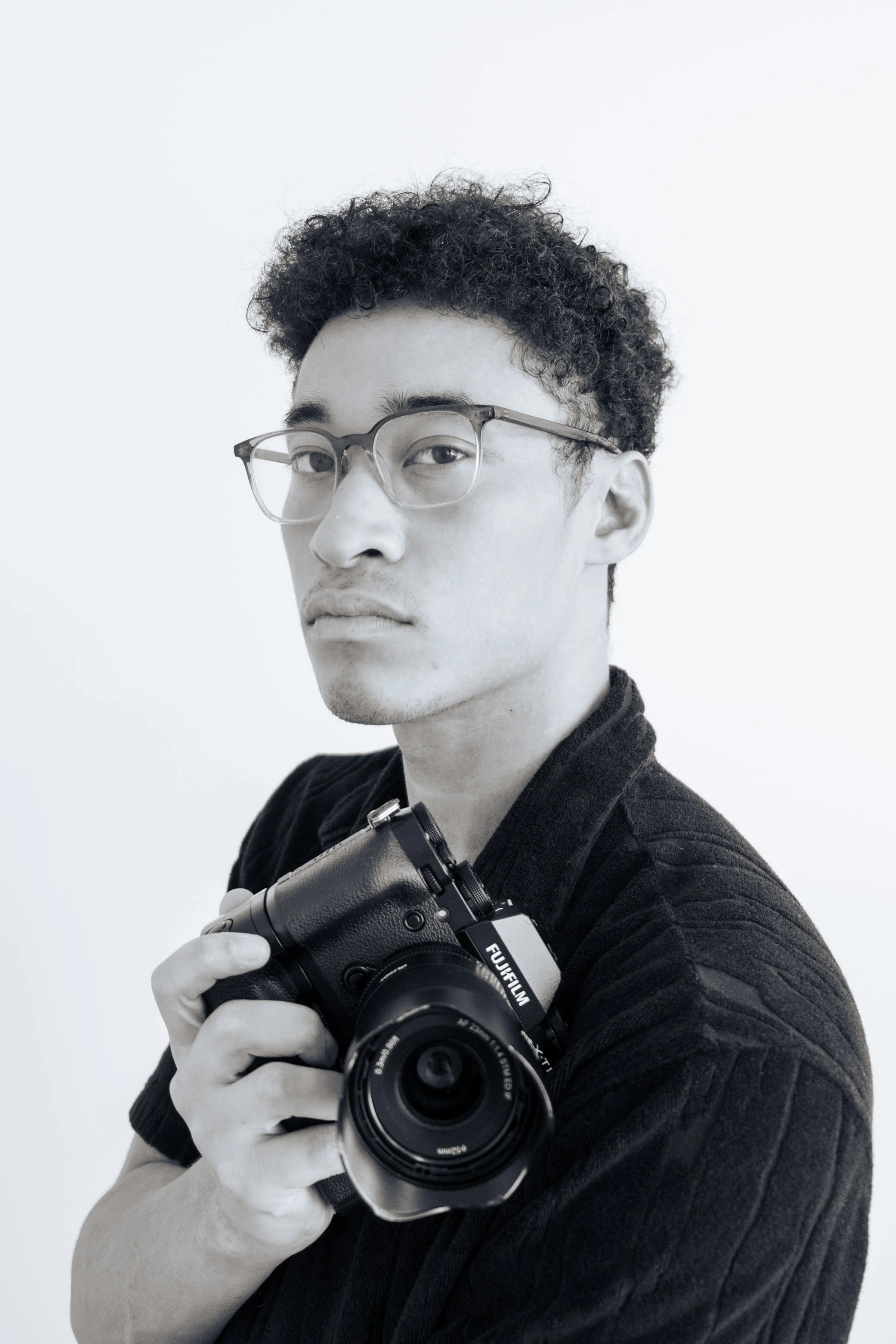 A person wearing glasses and a black blazer holding a black Fujifilm camera, taking a selfie against a plain white background.