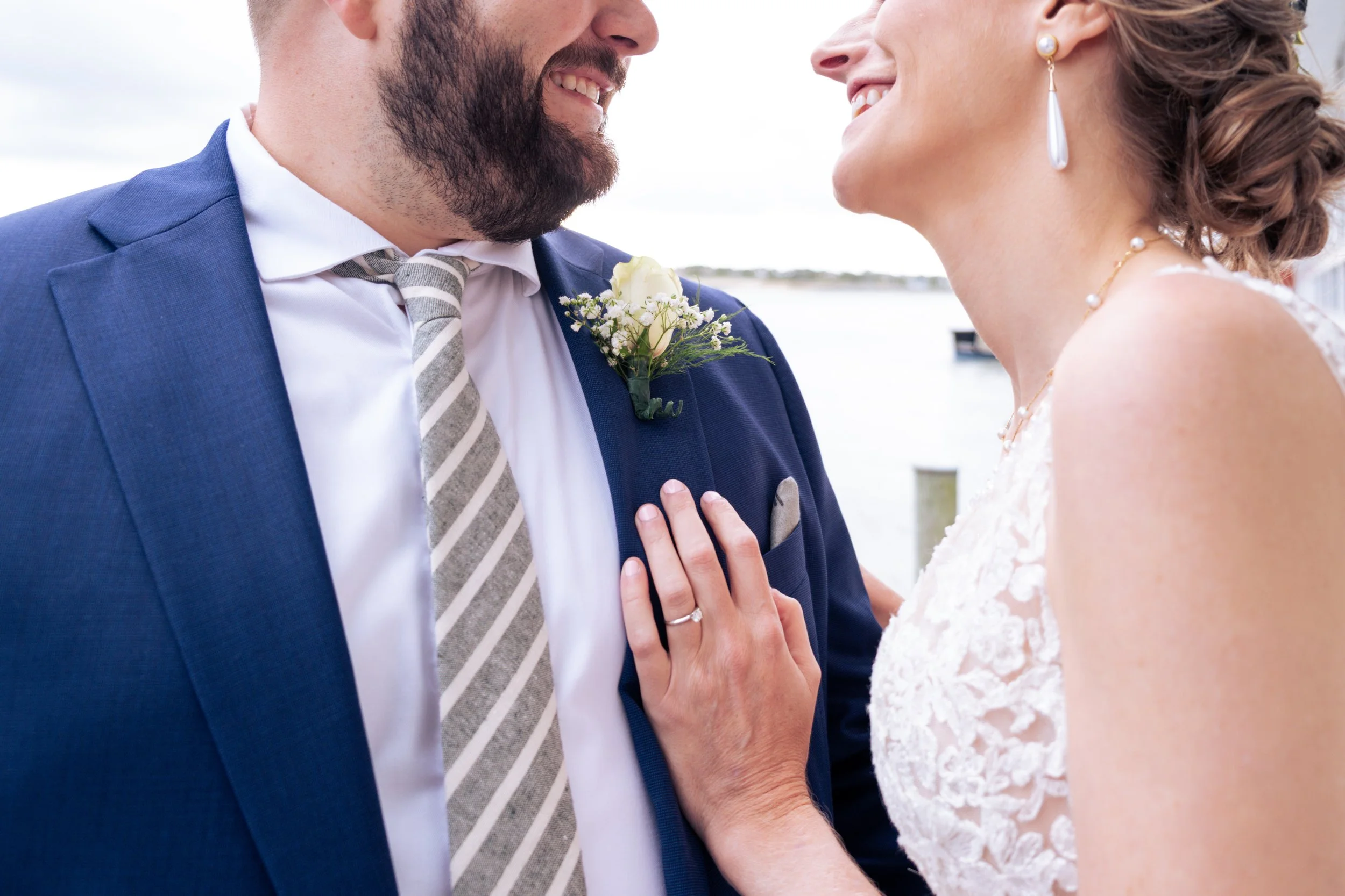 Close-up of a couple smiling at each other during their wedding. The groom wears a blue suit and a striped tie with a boutonniere, while the bride has pearl earrings, a necklace, and a white lace dress.