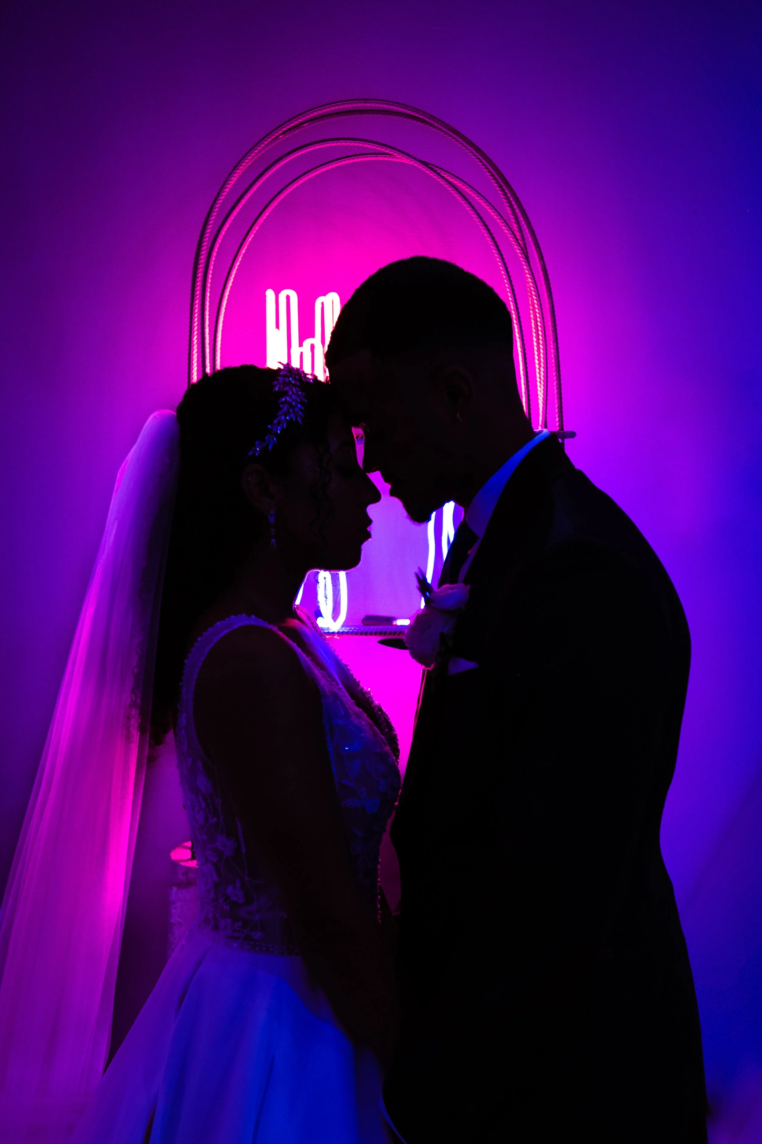 Silhouette of a bride and groom facing each other in front of a neon purple and pink background, with curved neon light decorations and a glowing neon sign behind them.