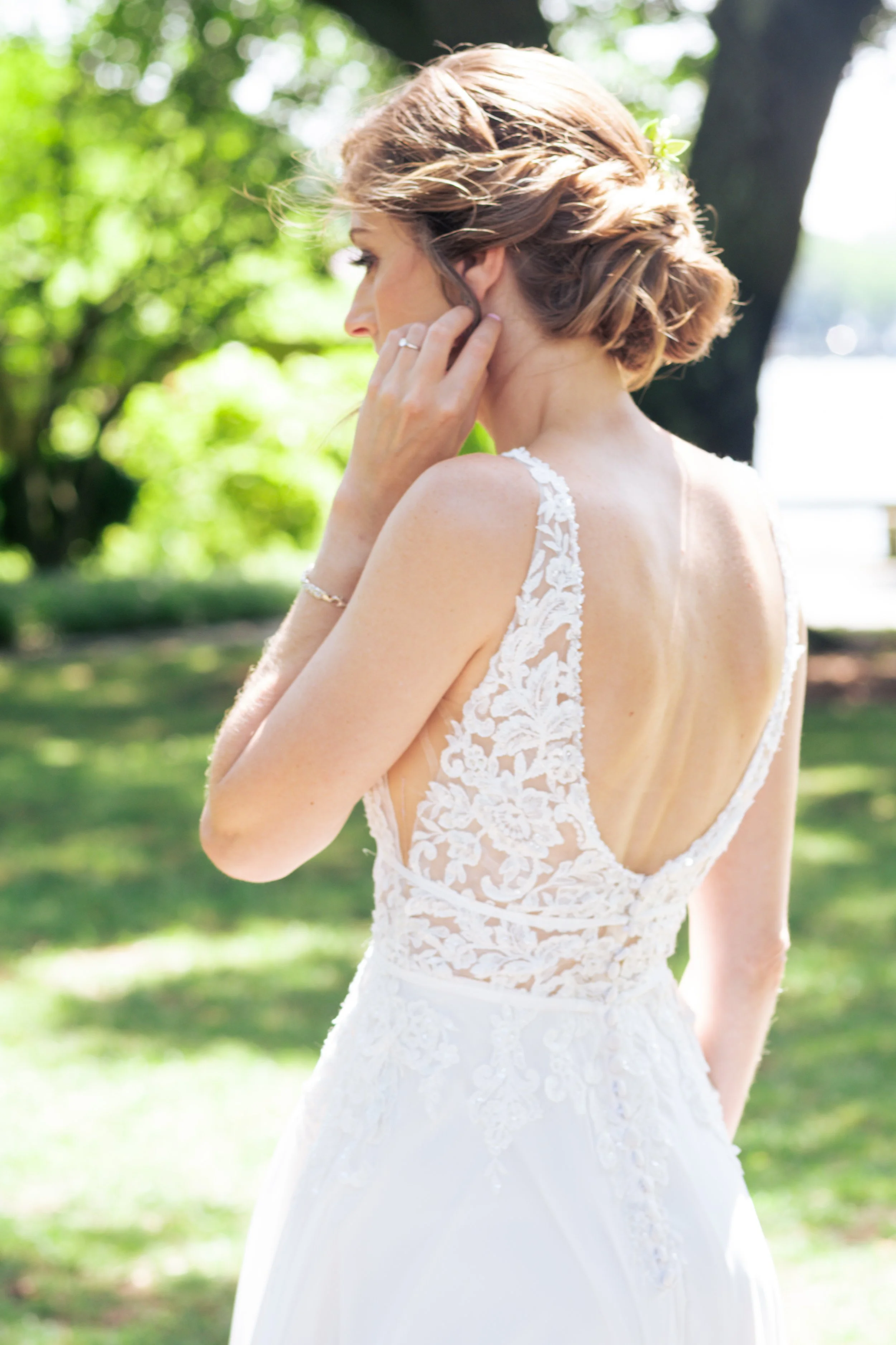 A woman with short, styled brown hair in a wedding dress outdoors with a tree and greenery in the background.