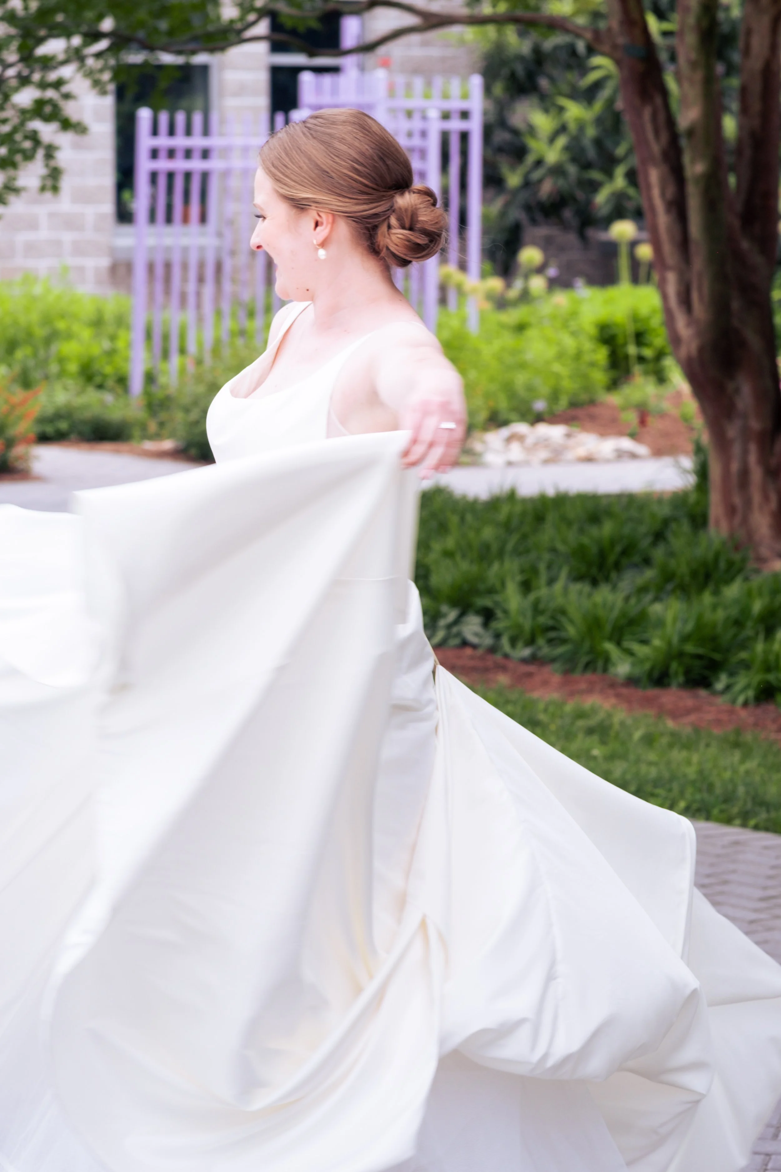 A woman dressed in a white wedding gown, smiling and twisting her gown in an outdoor garden setting with green plants, trees, and a brick building in the background.