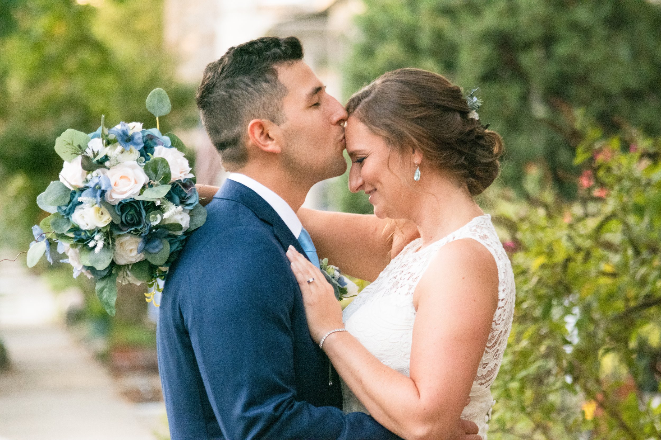 A bride and groom sharing a kiss during their wedding ceremony outdoors, with the bride holding a bouquet of white and blue flowers and greenery.