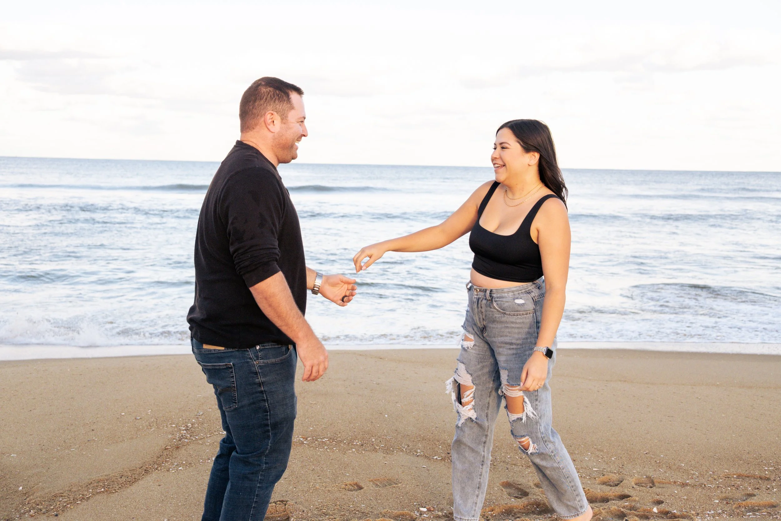 A man and woman are standing on the beach near the ocean, smiling and holding hands.