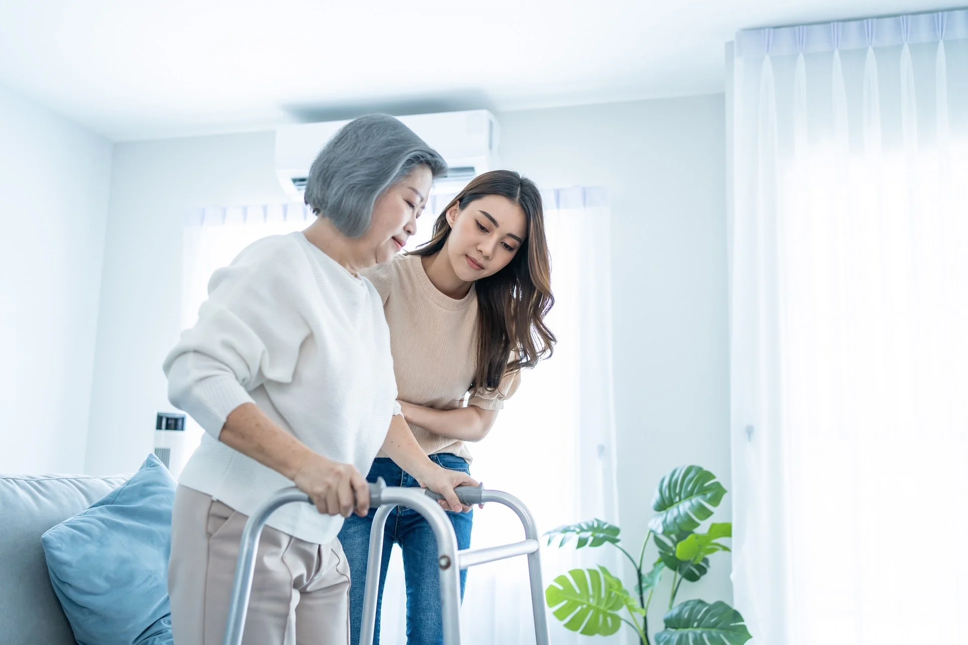 An elderly woman with gray hair using a walker is being assisted by a younger woman, possibly a caregiver or family member, in a bright, modern room with natural light and indoor plants.
