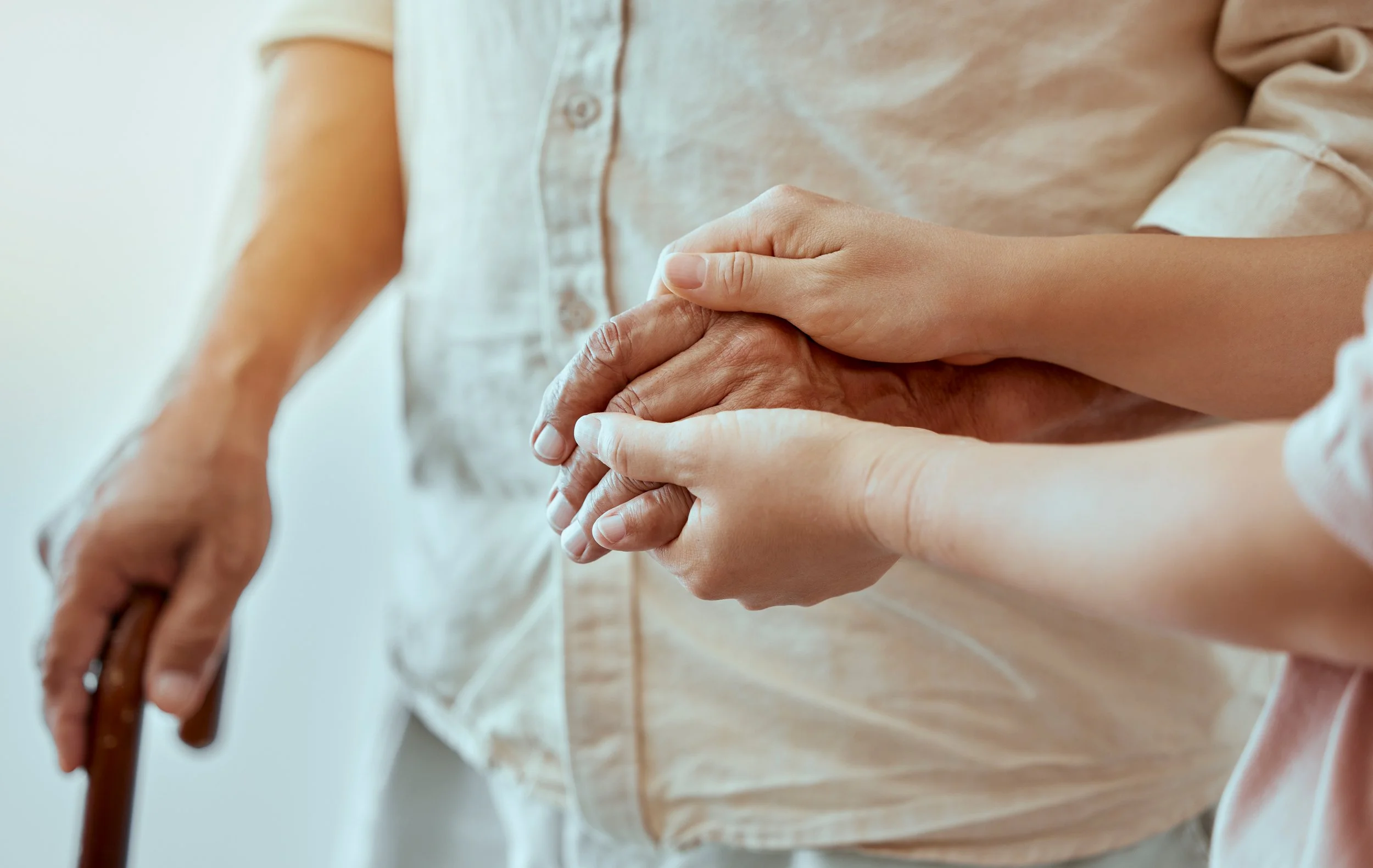Close-up of a person holding the hand of an elderly person, who is using a cane, with both hands gently clasped.