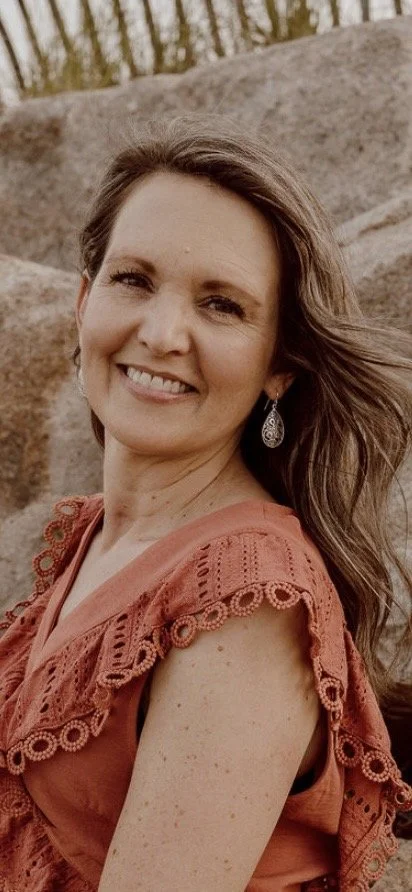 A smiling woman with long hair wearing orange top with lace trim, earrings, outdoors with bushes and rocks in the background.