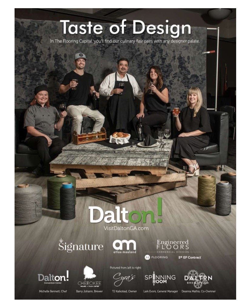 A group of five people, including two women in black dresses and a man in a chef's coat, sitting on black couches and standing behind a rustic wooden table, holding drinks. The background features a dark textured wall and construction items like spools of thread on the floor. The image promotes Dalton and its culinary and flooring partners.