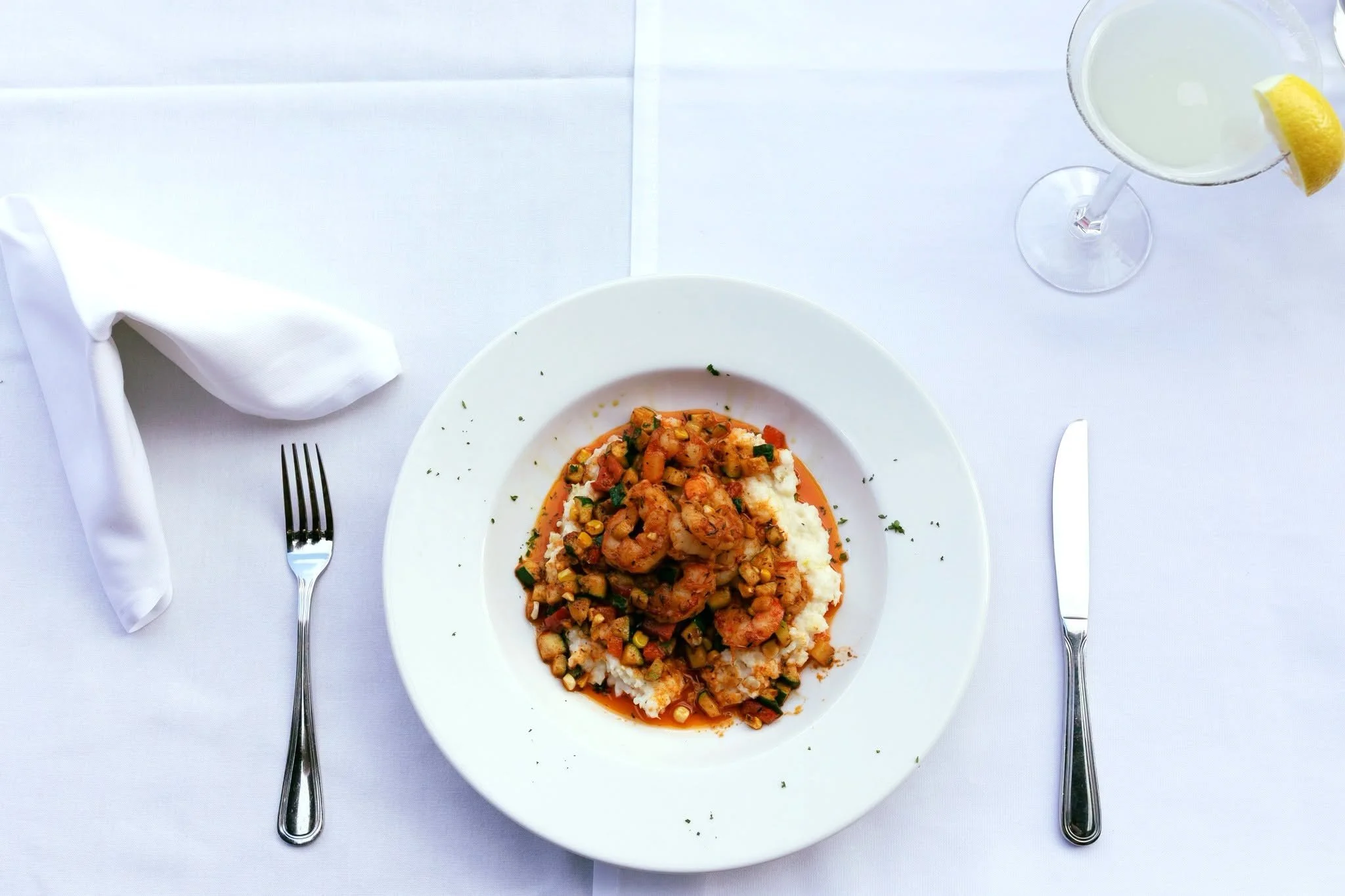 White plate with shrimp and vegetable dish served with white mashed potatoes and gravy, set on a white tablecloth with a fork on the left and a knife on the right, a rolled napkin and a lemon garnish in a cocktail glass.