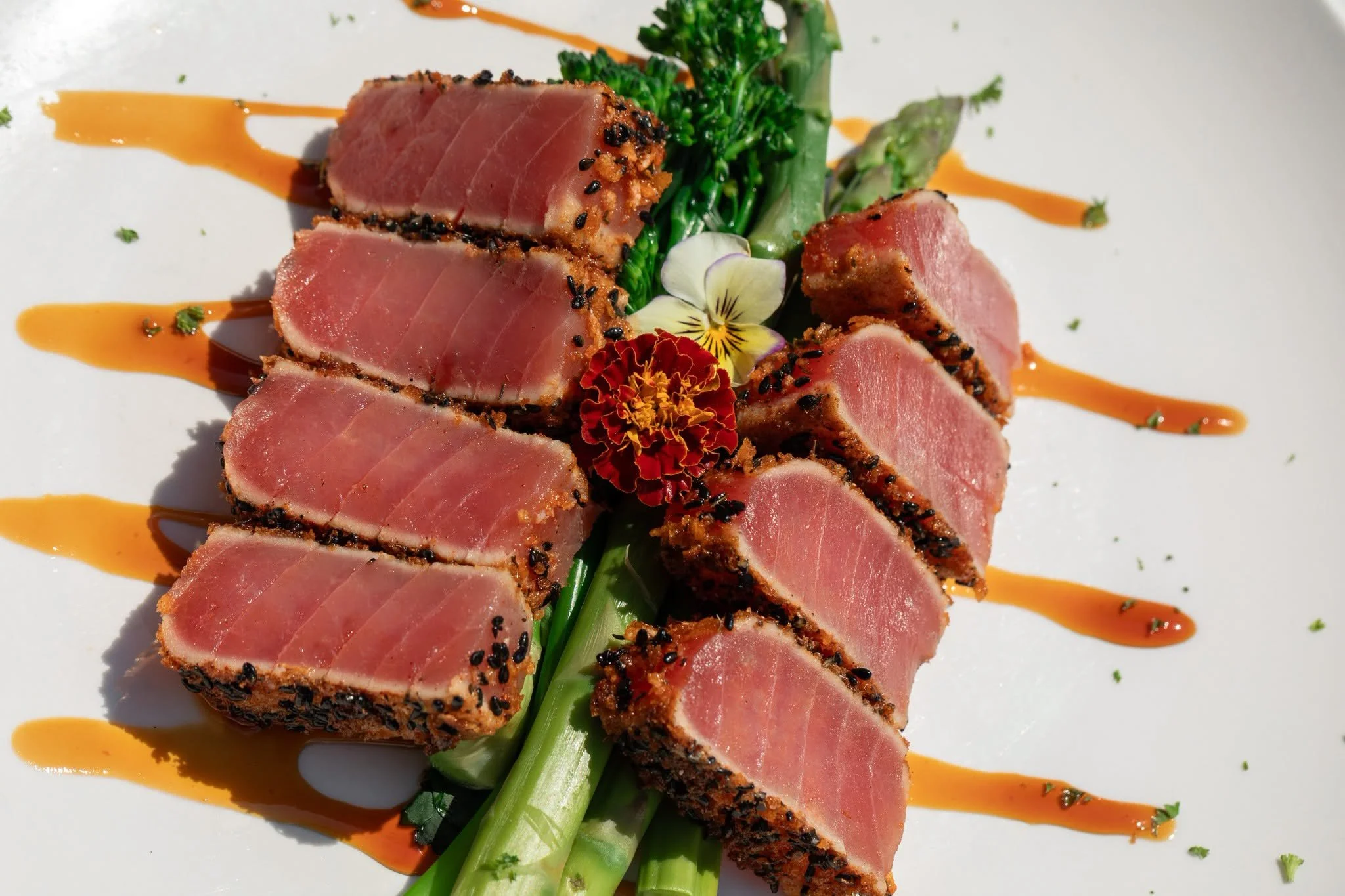 Slices of seared tuna coated with black sesame seeds, garnished with green vegetables, edible flowers, and drizzled with orange sauce on a white plate.
