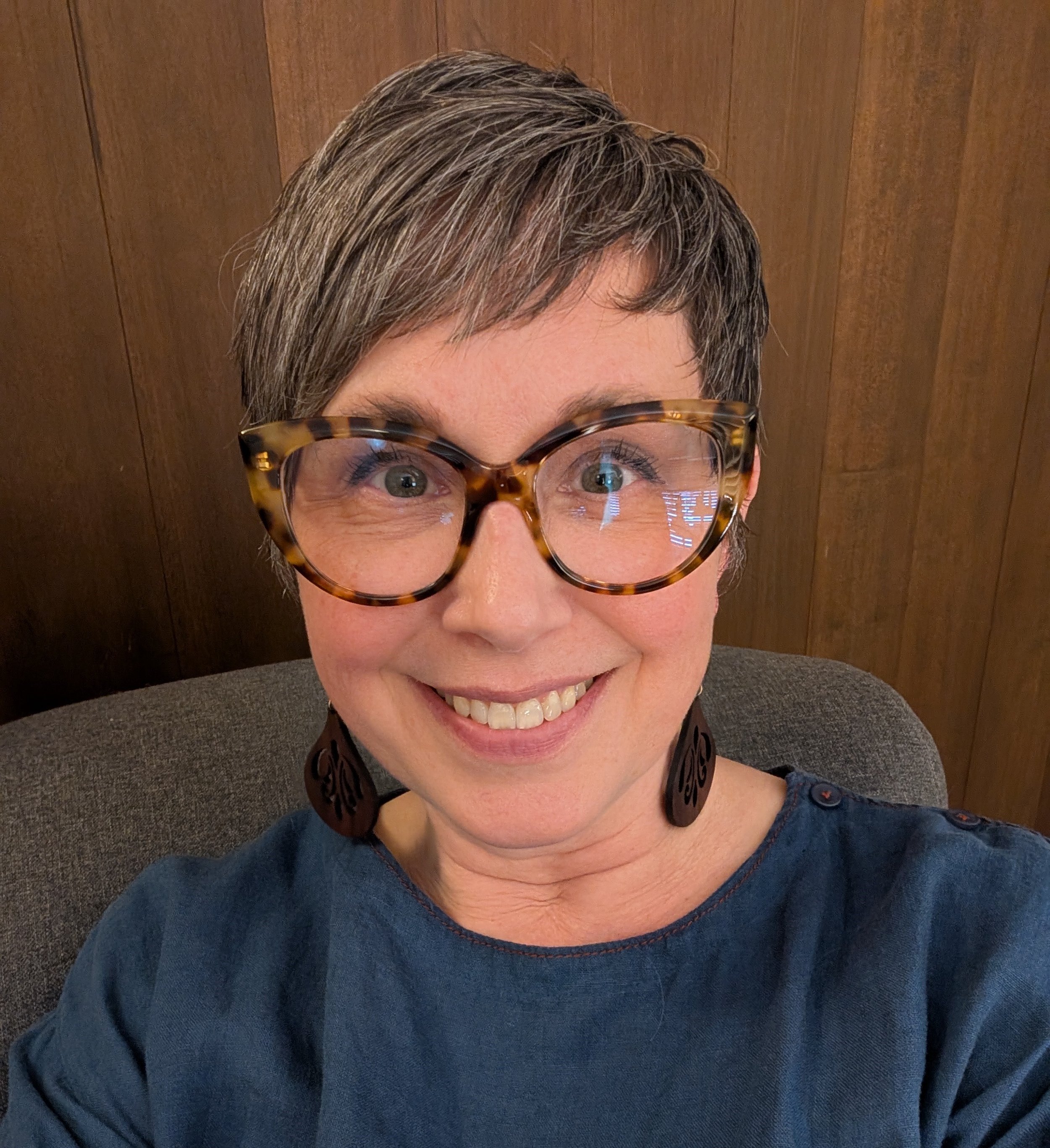 Close-up of a woman with short gray hair wearing tortoise-shell glasses, smiling, against a wood-paneled background.