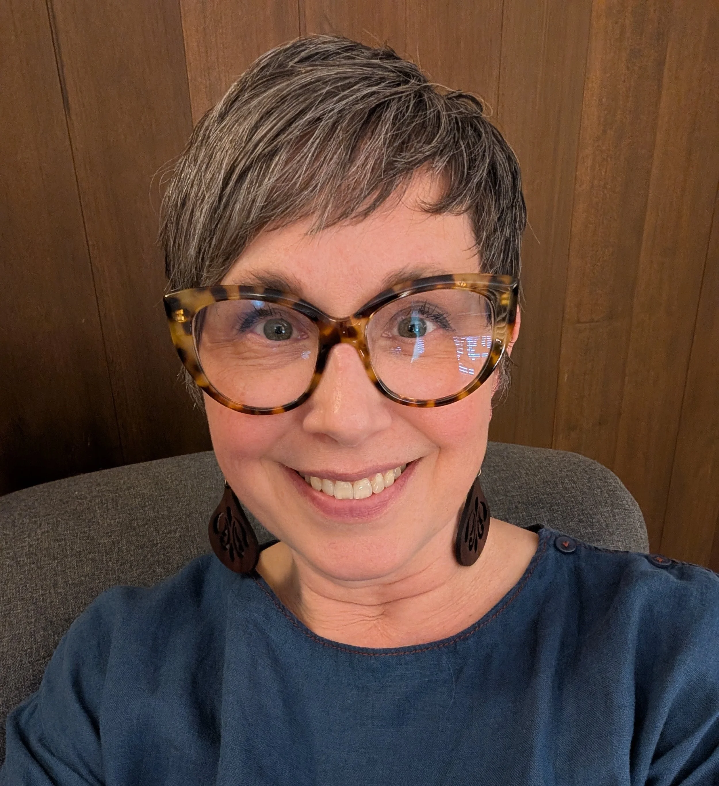Close-up of a woman with short, gray hair wearing tortoiseshell glasses and a dark blue shirt, smiling while sitting with a wooden wall in the background.