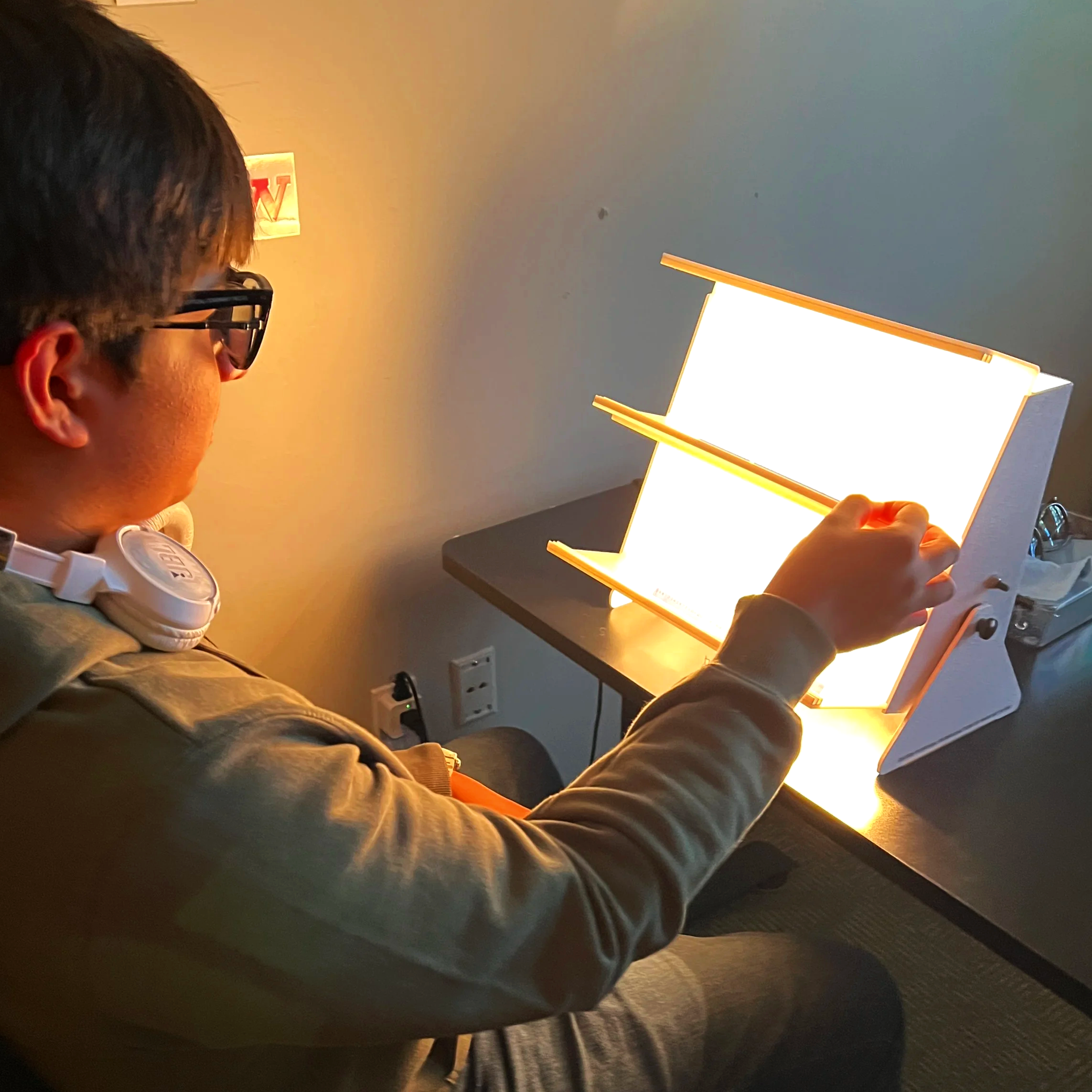 A person with dark hair and glasses sitting at a desk, holding a tablet with a bright screen. There is a lamp on the desk and a power outlet on the wall behind them.