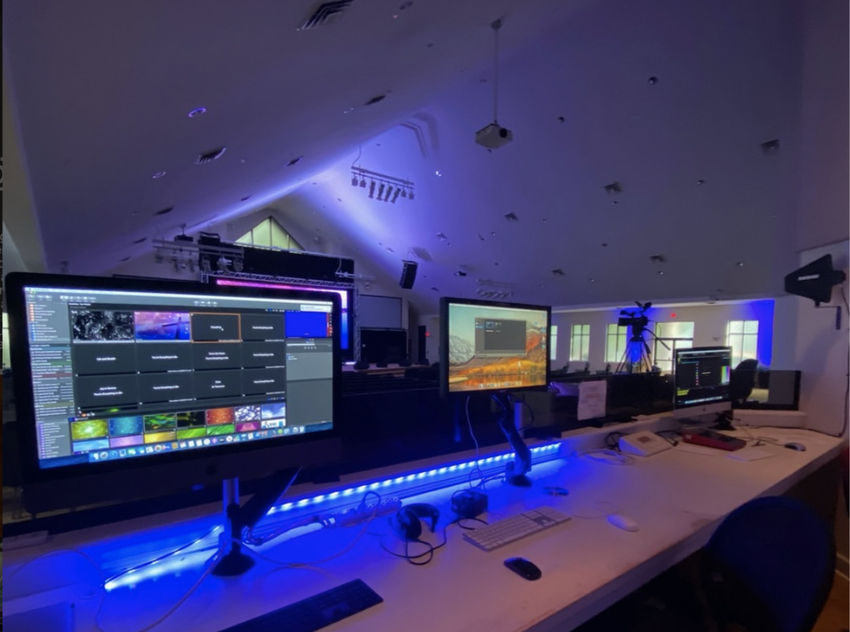 A control room with multiple computer monitors, some displaying video editing software and colorful visuals, illuminated by blue LED lights, with stage lighting and cameras set up in the background.