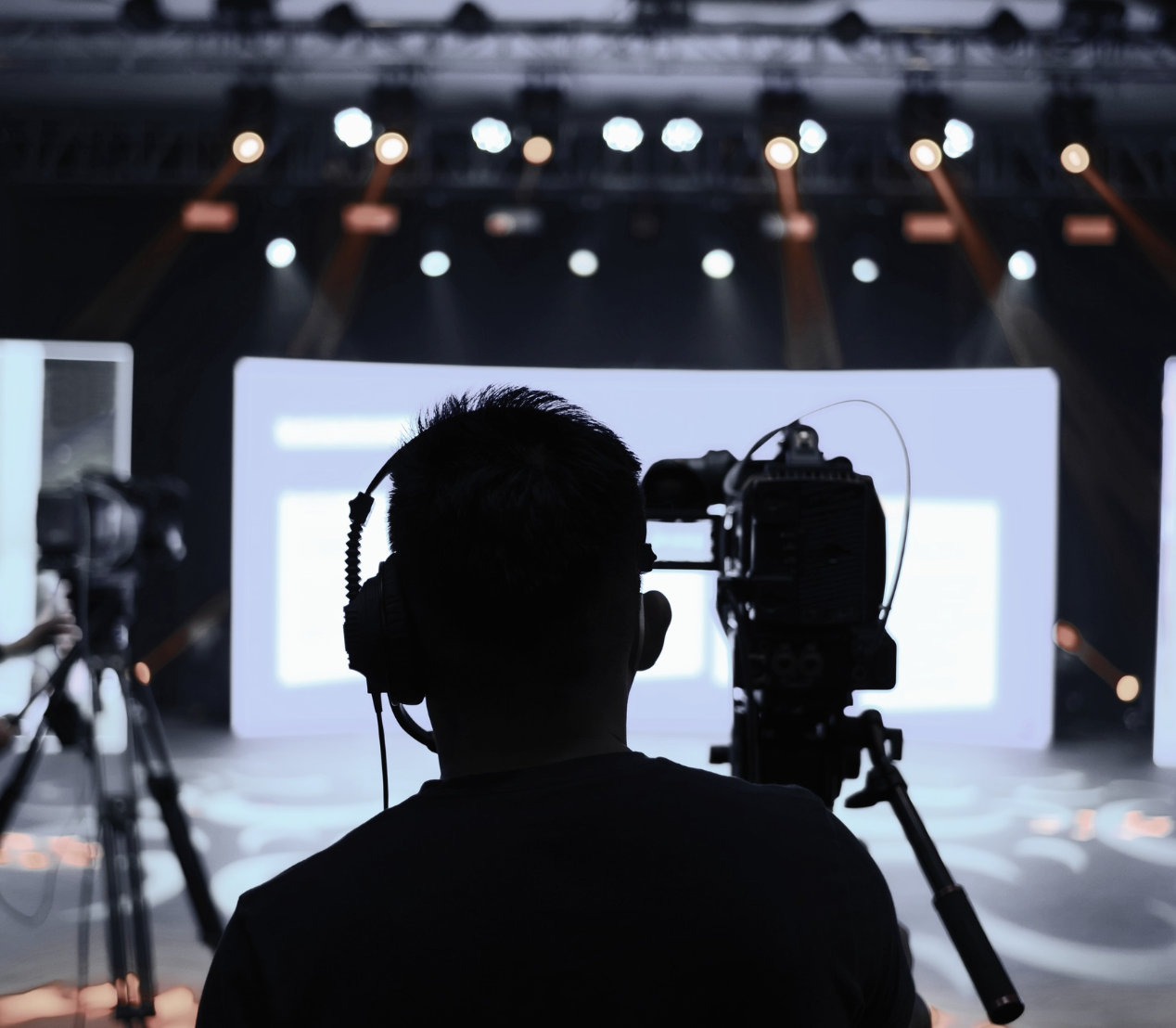 Silhouette of a person with headphones operating a video camera at a conference or event with a large screen and stage lights in the background.