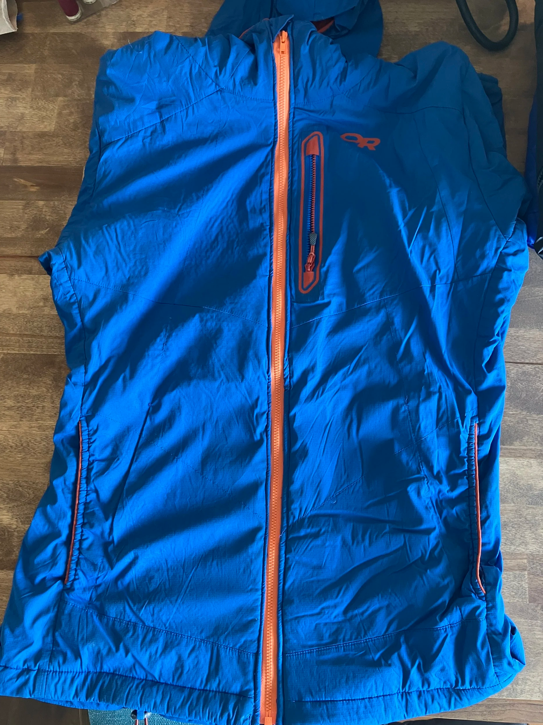 Blue outdoor jacket with orange zipper and logo, laid flat on a wooden surface.