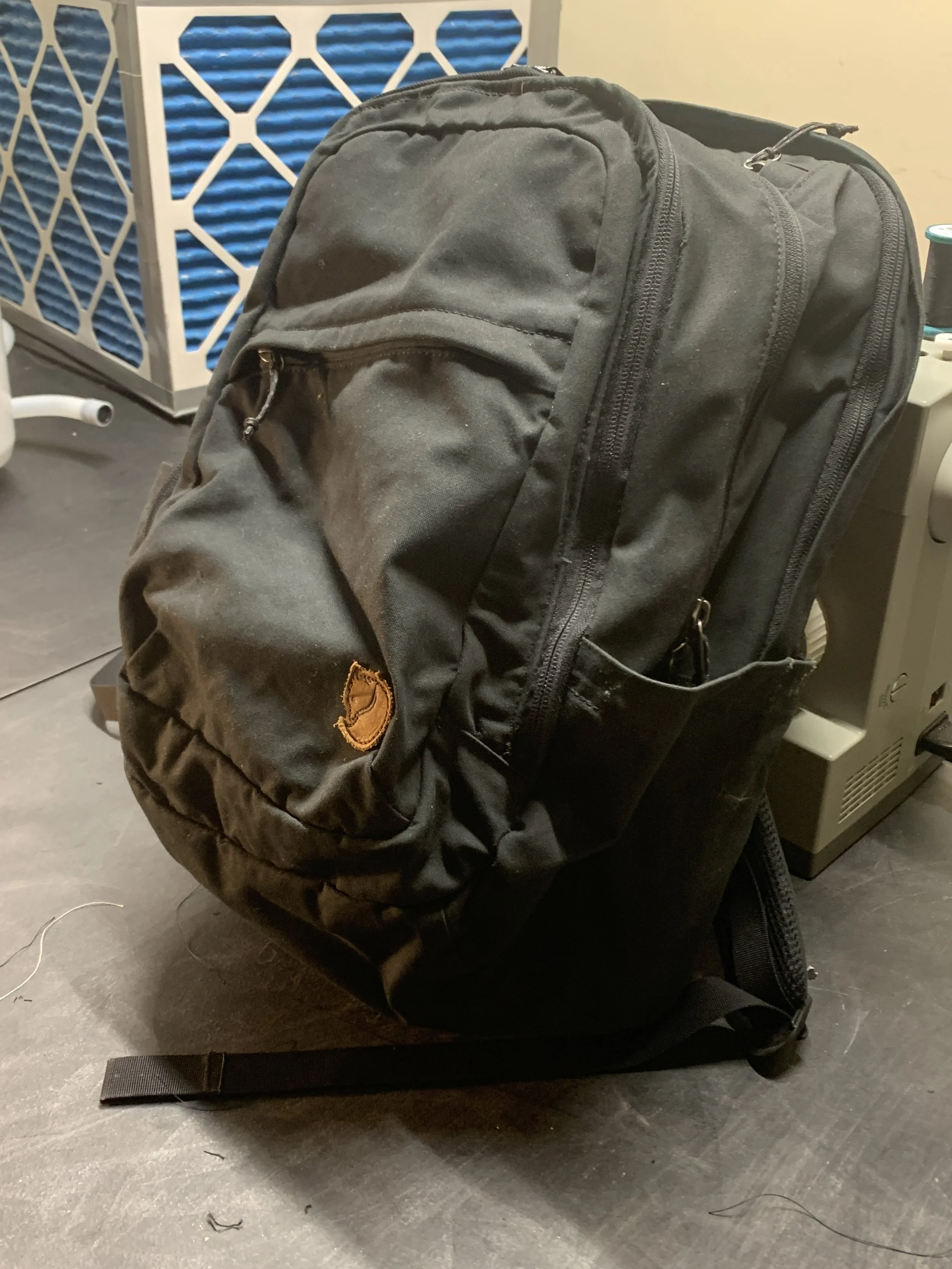 Black backpack with multiple zippered compartments resting on the floor next to a piece of office equipment and a blue vented panel.