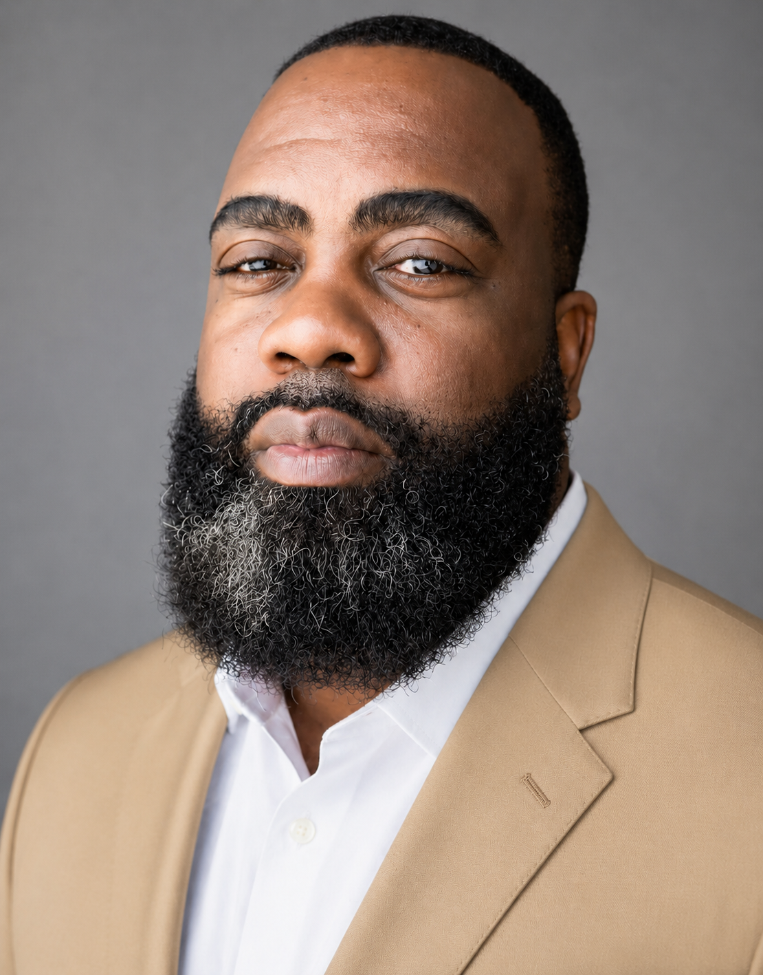 Close-up portrait of a confident African American man with a full beard and short hair, wearing a beige suit jacket and a white shirt, against a plain gray background.