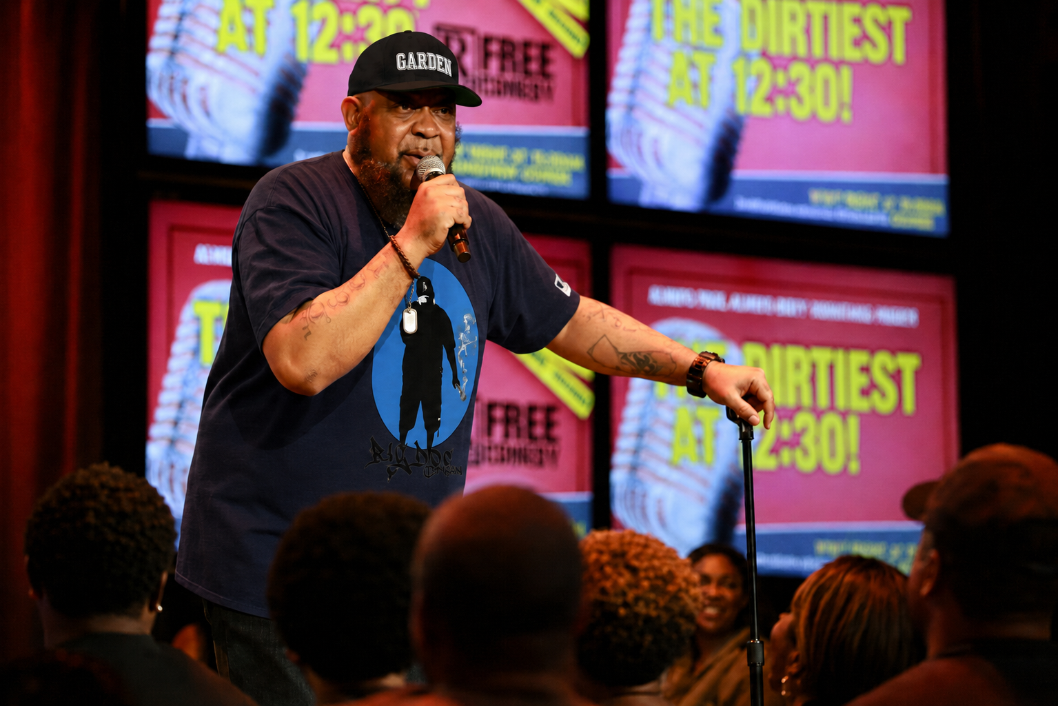 A man with a beard and tattoos, wearing a black cap and a black t-shirt, speaks into a microphone on stage, in front of a screen that shows a colorful poster for an event called 'The Dirtiest at 12:30!'. The audience is visible in the foreground.
