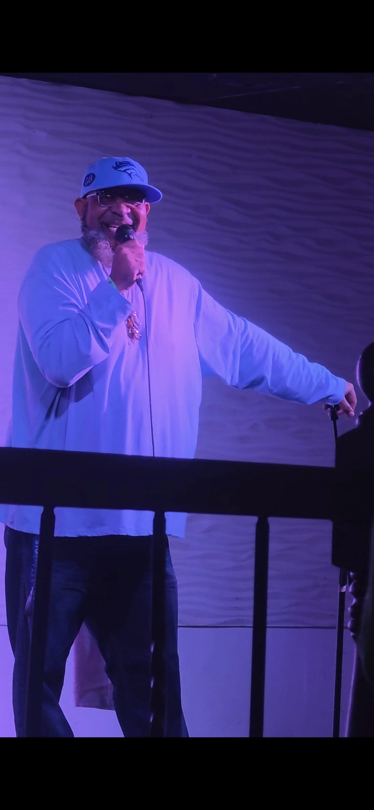 Man wearing a Denver Broncos cap and glasses, holding a microphone, smiling on stage with purple lighting.
