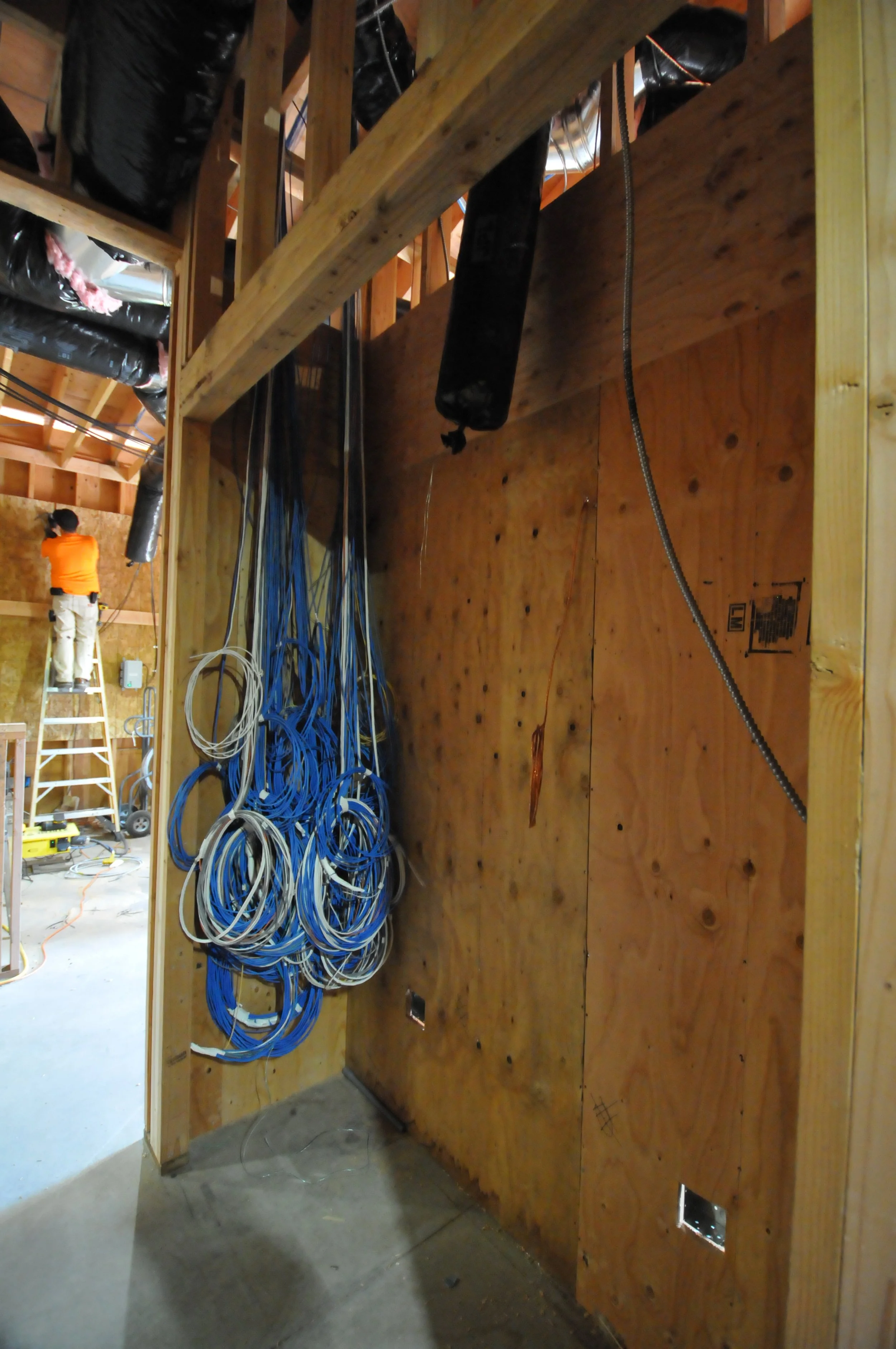 A construction site with a partially built room, showing wooden studs, electrical wiring, and ductwork. A man in an orange shirt is standing on a ladder working on electrical wiring in the background.