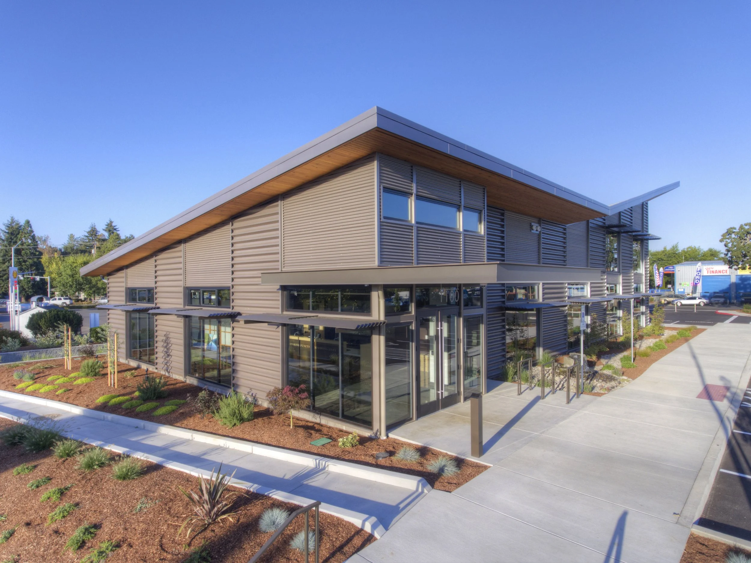 Modern commercial building with large glass windows, metal siding, and a landscaped area with small plants in front.