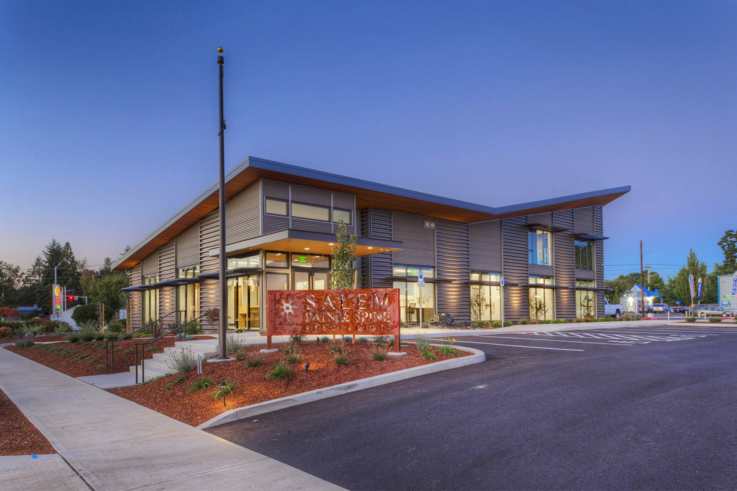 Modern two-story building with large windows, wooden accents, and a sloped roof. There is a parking lot with empty spaces, a sign for Salem Pain and Spine Specialists, and landscaped area with small plants and mulch. The sky is clear with a fading su