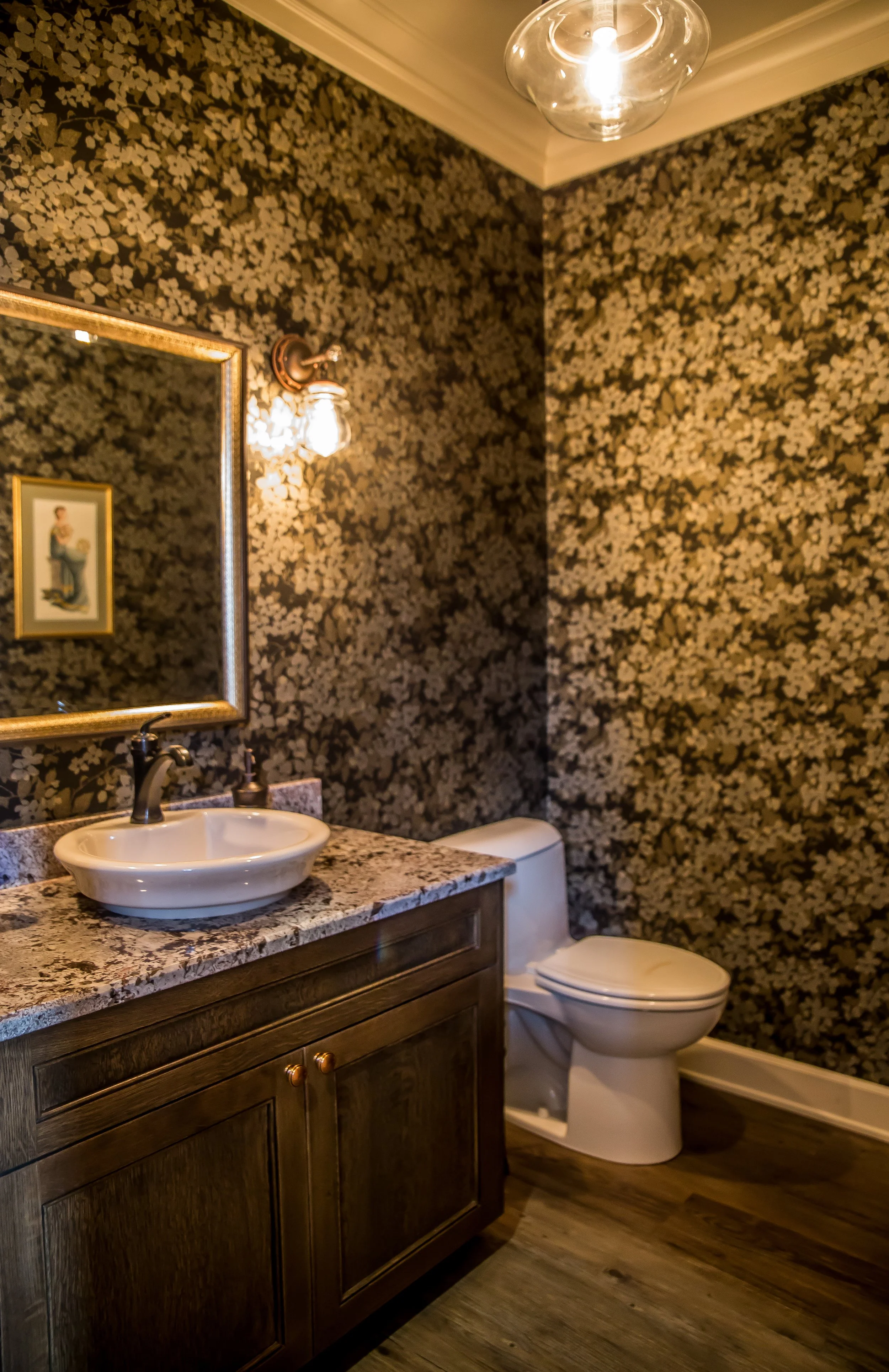 Bathroom with floral wallpaper, a wooden vanity with a granite countertop, a vessel sink, a framed mirror, a wall sconce, a toilet, and a wooden floor.