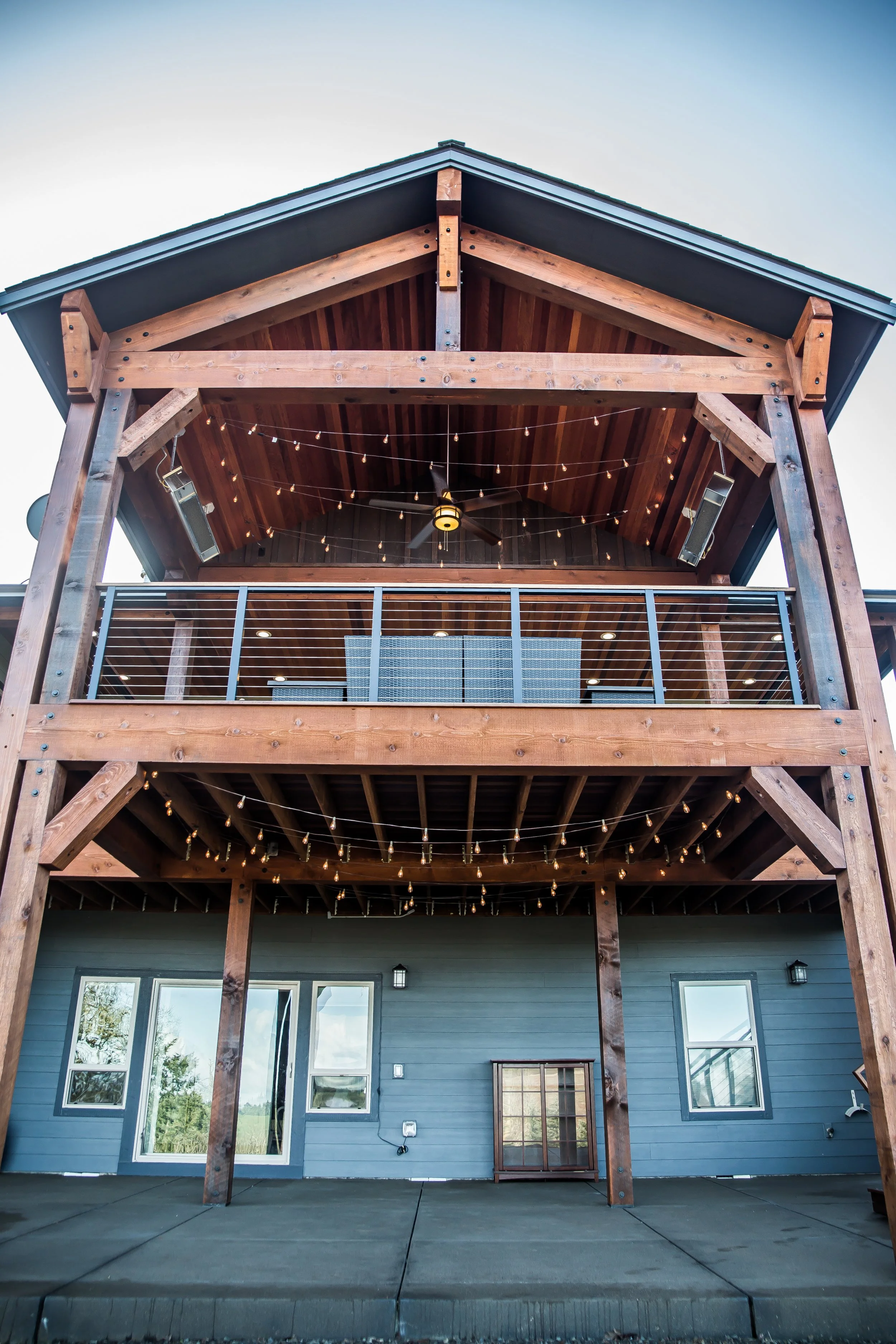 The image shows the exterior of a two-story house with a wooden balcony on the upper level. The balcony has a wooden railing, string lights, and a ceiling fan. The house has gray siding, multiple windows, and a concrete patio area below the balcony.