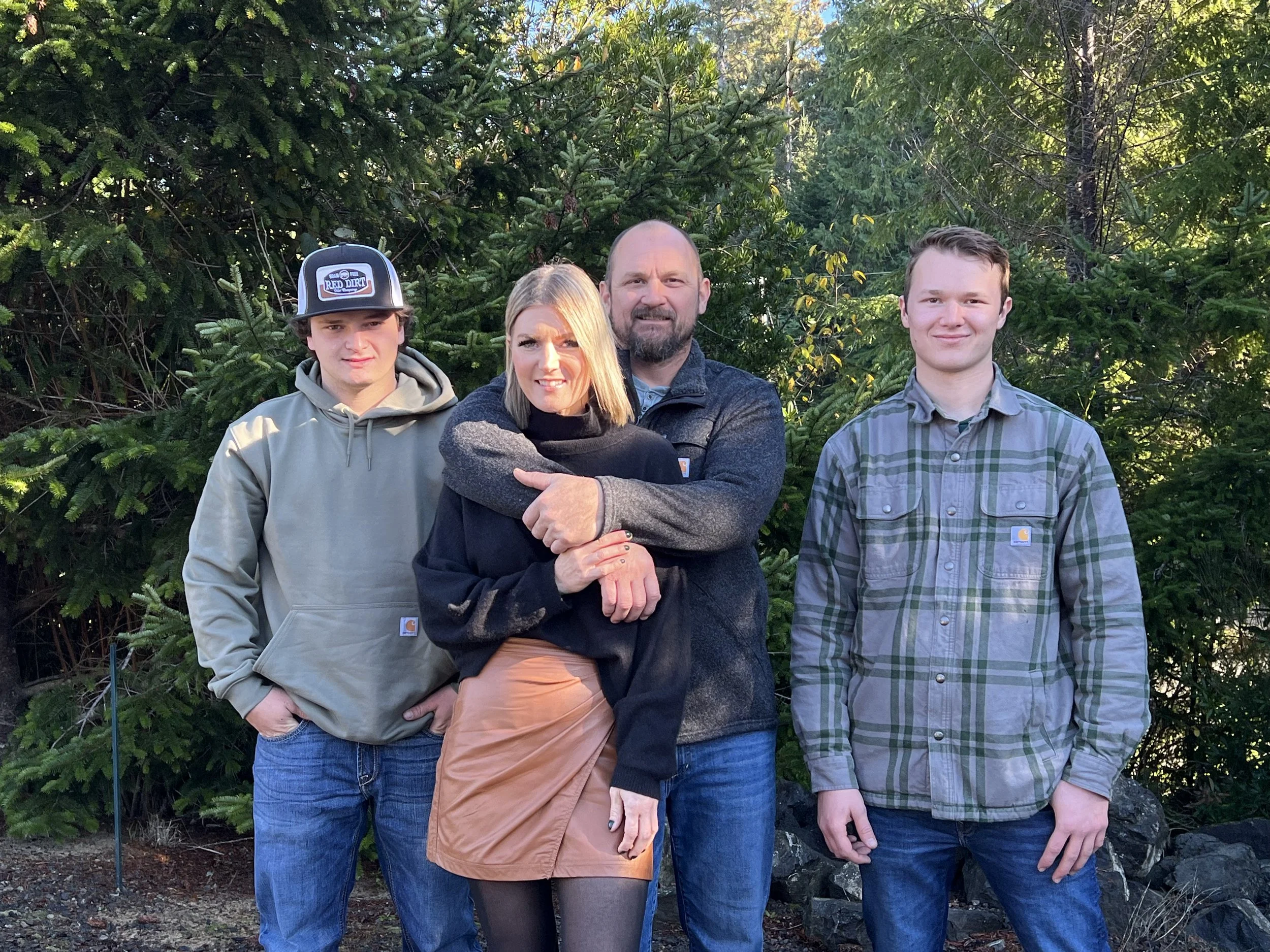 A family of four standing outdoors in front of green trees. The woman is embracing the man, and two young men are standing beside them, all smiling.