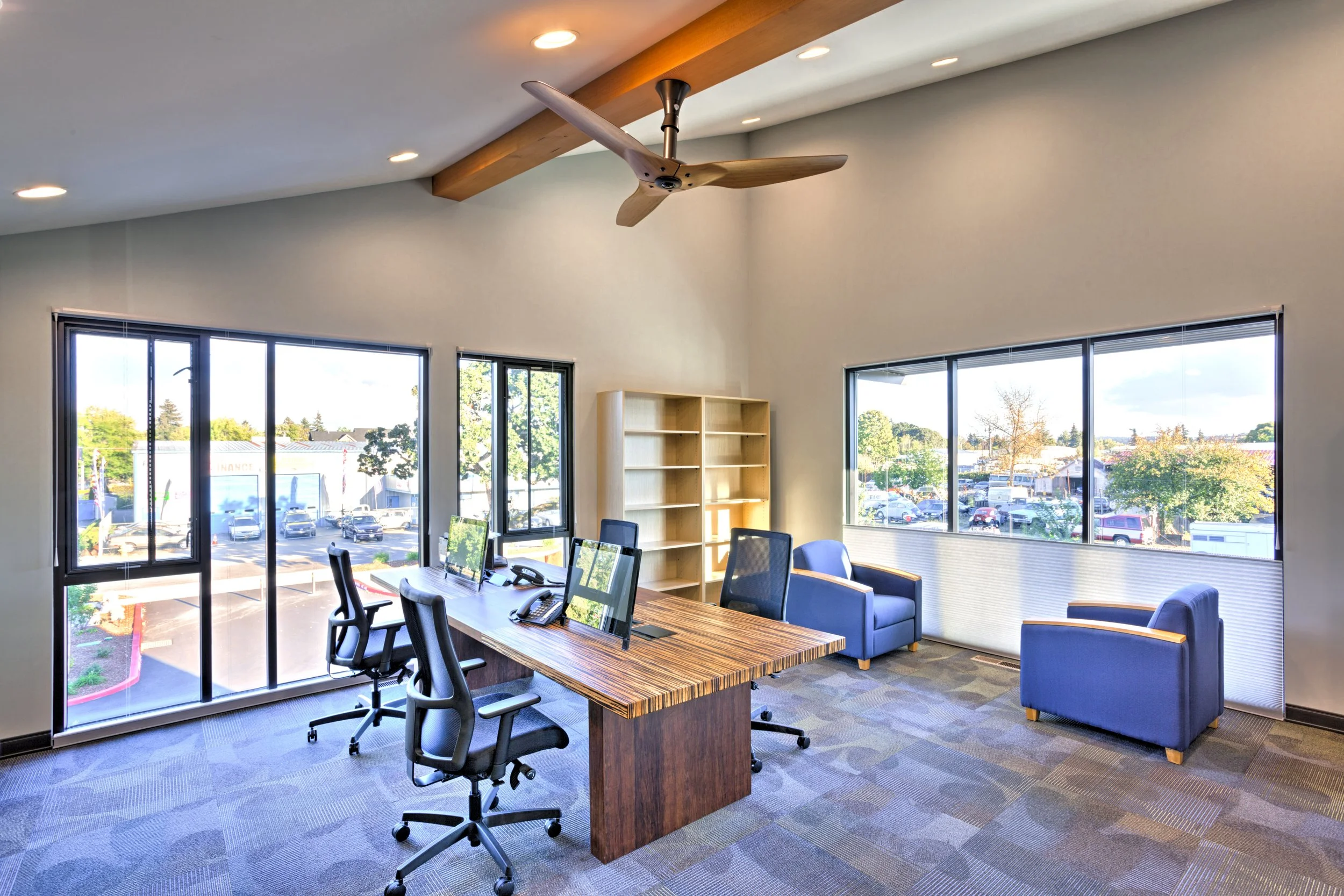 Office conference room with wooden table, multiple black office chairs, blue armchairs, large windows, ceiling fan, ceiling lights, outside parking lot and trees visible