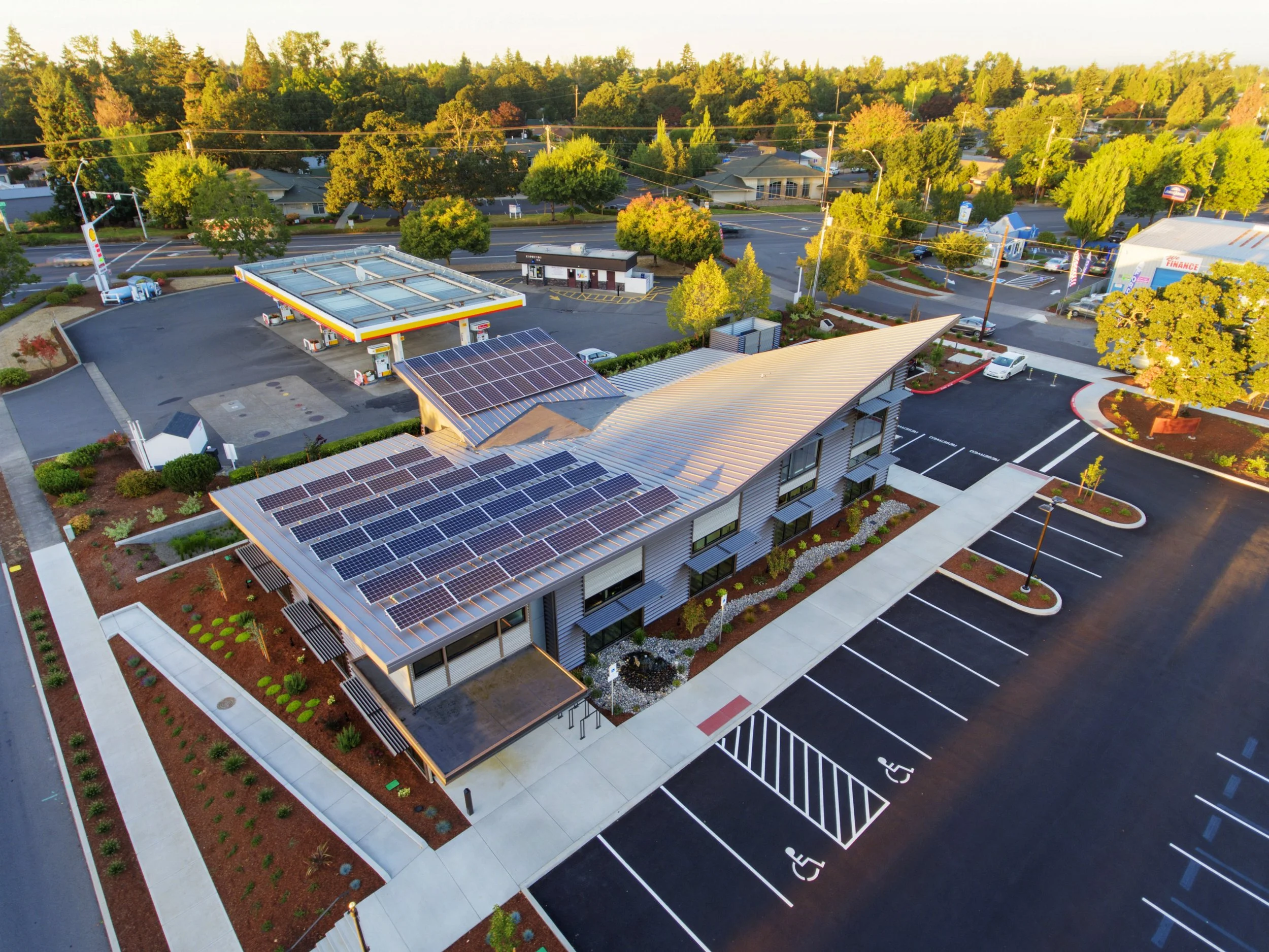 An aerial view of a modern building with solar panels on the roof, surrounded by parking lot spaces including designated accessible parking spots, and a gas station nearby, with trees and residential houses in the background.