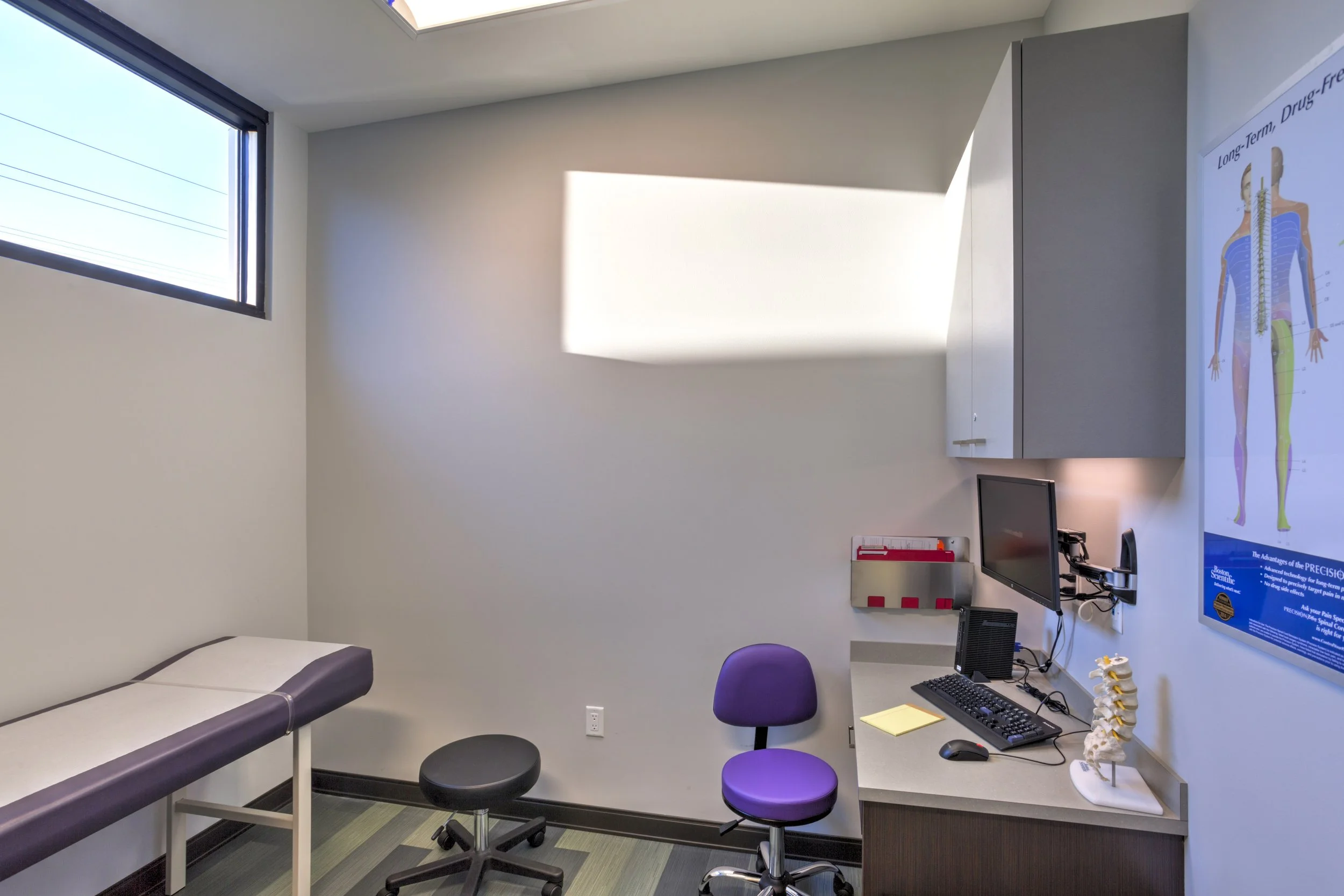 Medical examination room with a patient bed on the left, a computer monitor, and an educational poster about long-term drug therapy on the right wall. There are two stools, a cabinet, and a model of the human spine on the desk.
