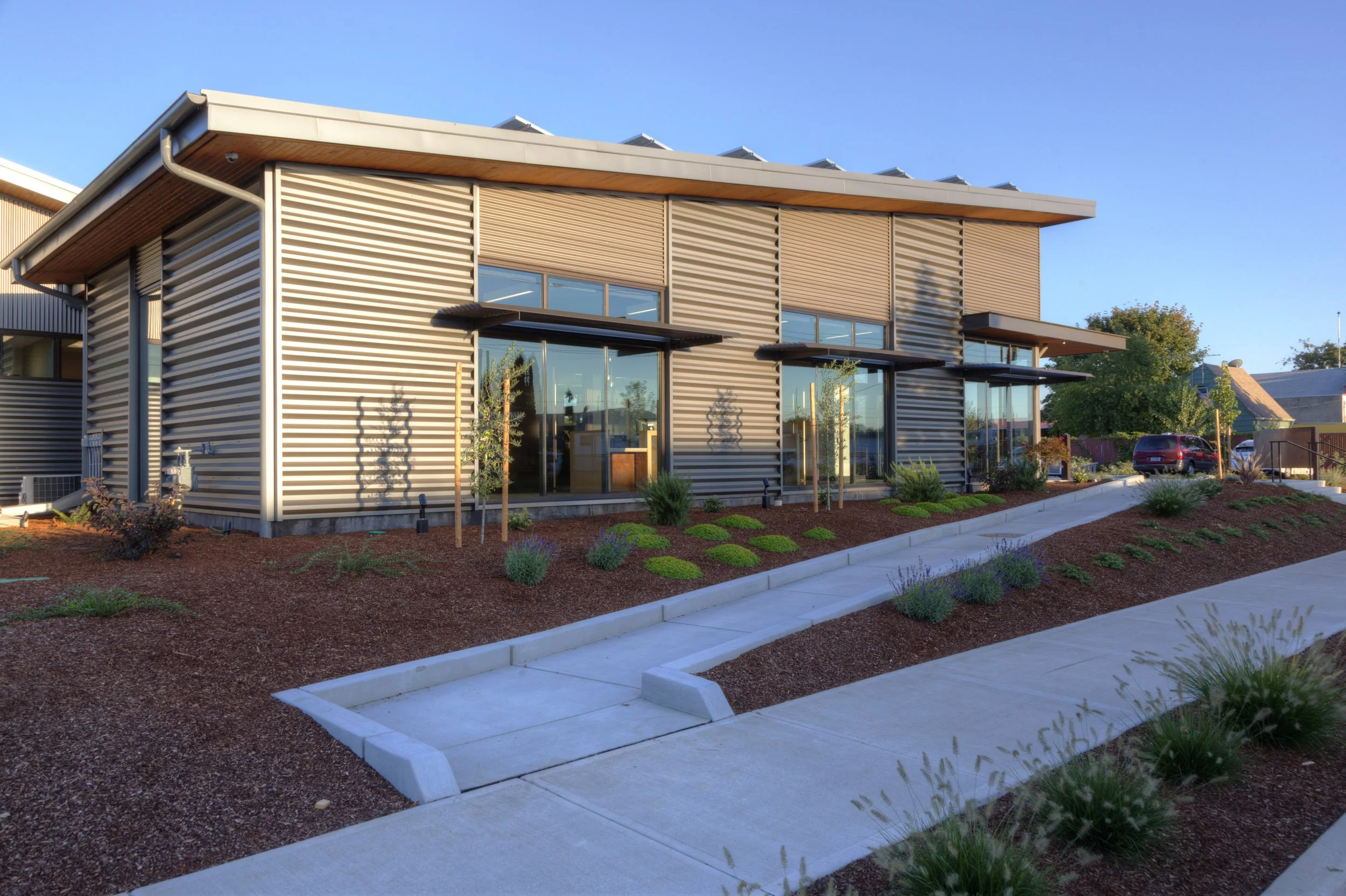 Modern commercial building with metal siding, large glass windows, and landscaped foreground with small plants and a paved walkway.